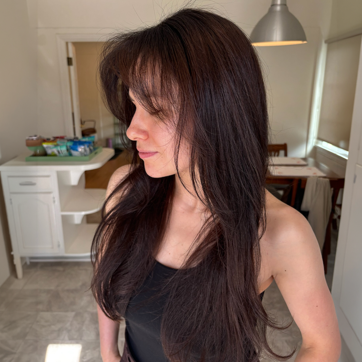 A woman with long dark brown hair, wearing a black sleeveless top, standing in a room with a kitchen and dining area in the background.