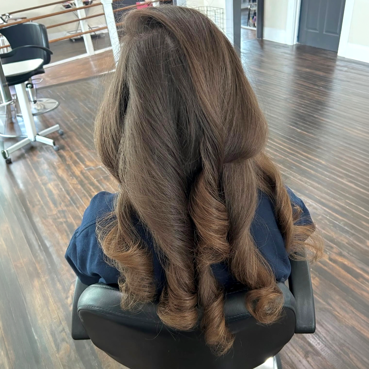 Back view of a woman with long, brown hair styled in loose curls, sitting in a salon chair inside a hair salon.