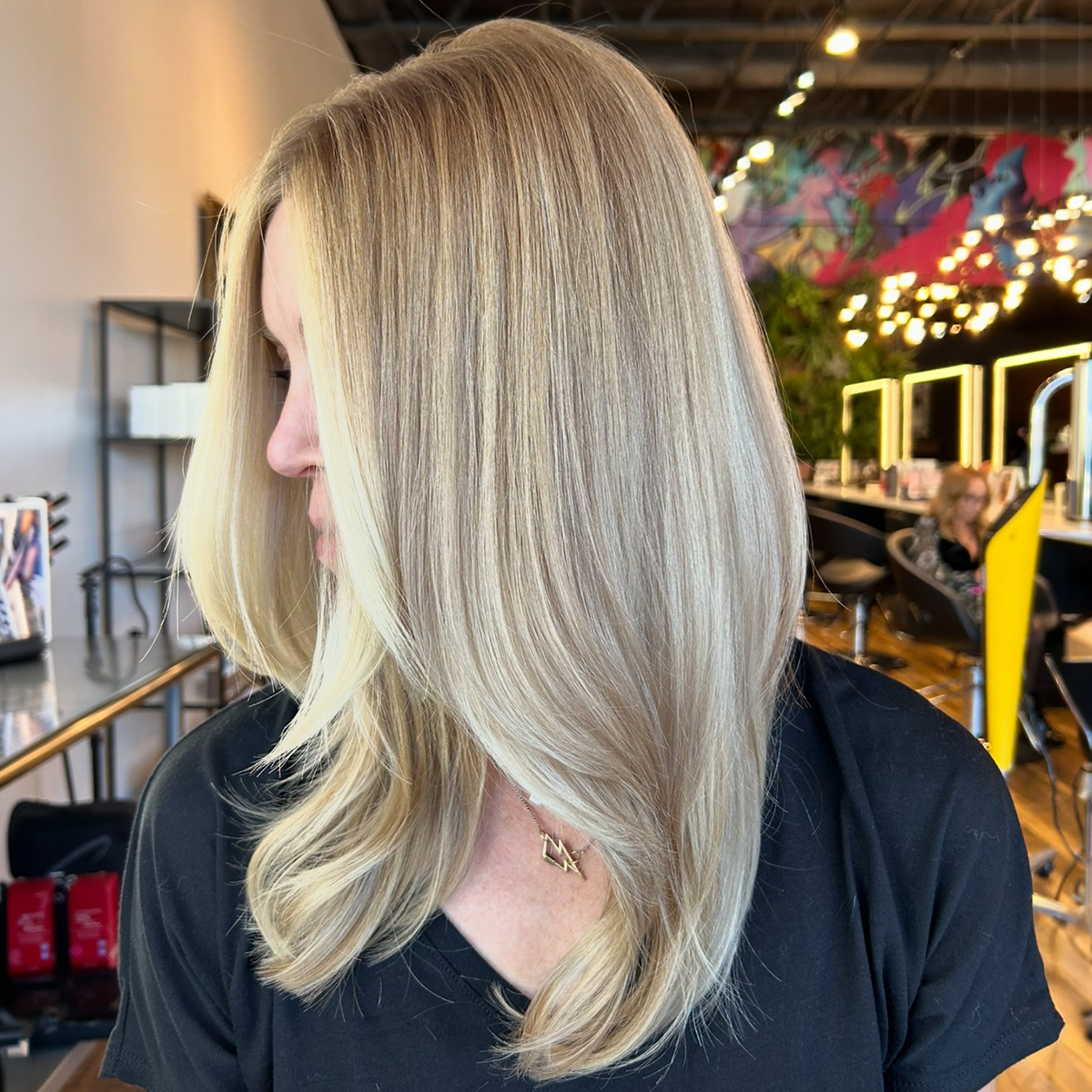 Woman with blonde, wavy hair in a salon with curved mirrors and colorful wall art.