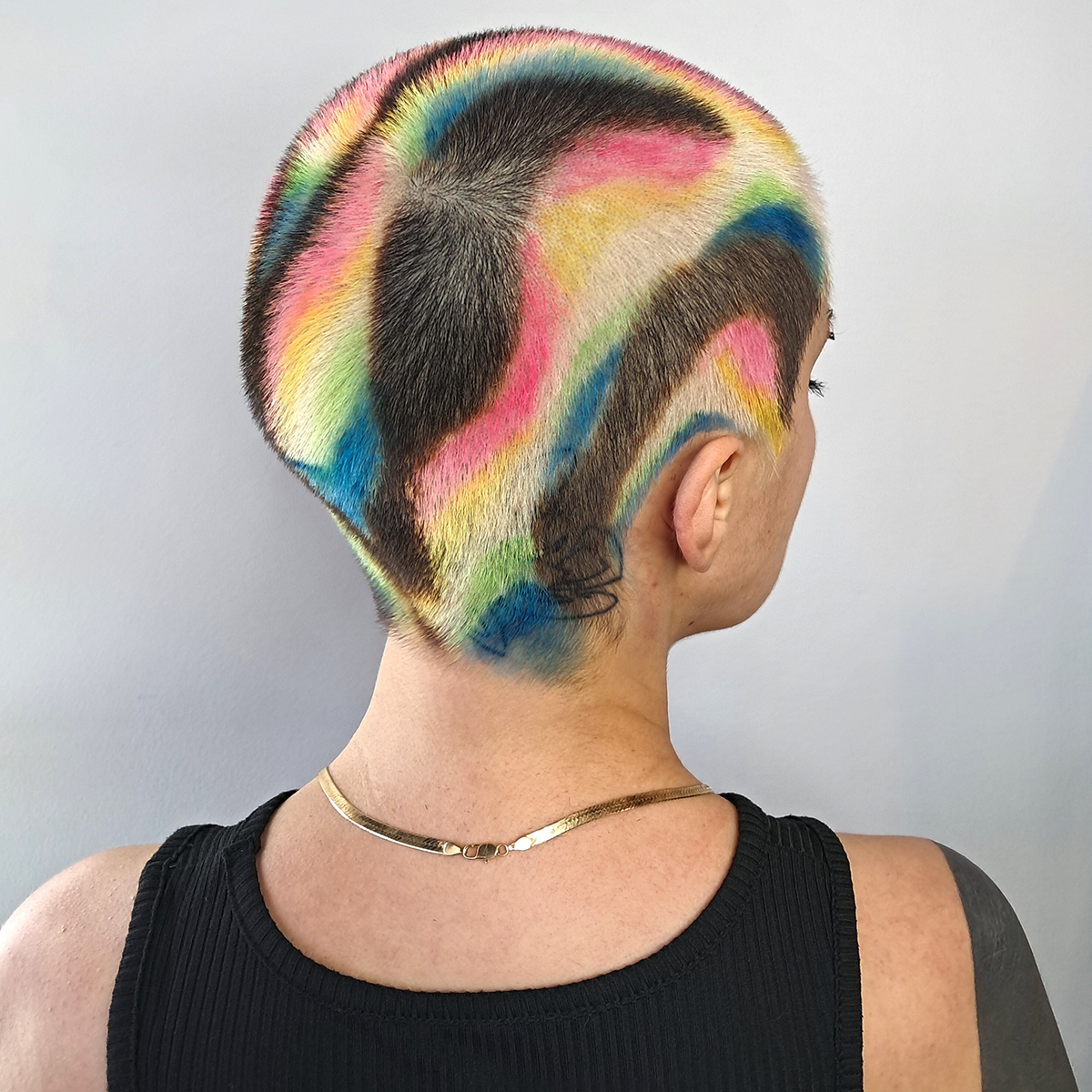 Side view of a person with short hair painted in colorful, abstract patterns including pink, yellow, blue, green, and black, on a plain background.
