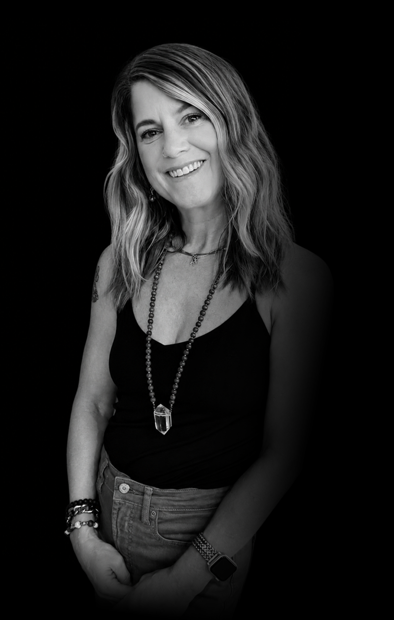 Black and white portrait of a smiling woman with wavy hair, wearing a necklace, bracelets, a black sleeveless top, jeans, and an Apple Watch, against a black background.