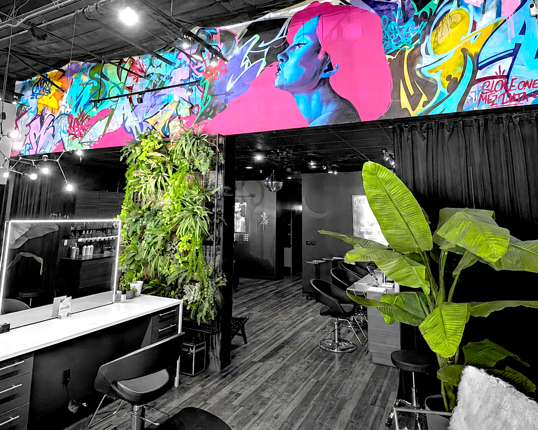 Interior of a modern hair salon with black walls, large green plants, and colorful graffiti art of a woman's face with pink hair on the wall above.