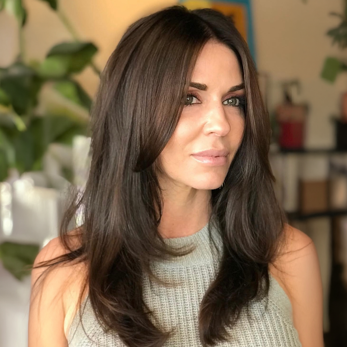 A woman with long, dark brown hair and a light makeup look, wearing a sleeveless knitted top, in an indoor setting with plants and shelves in the background.