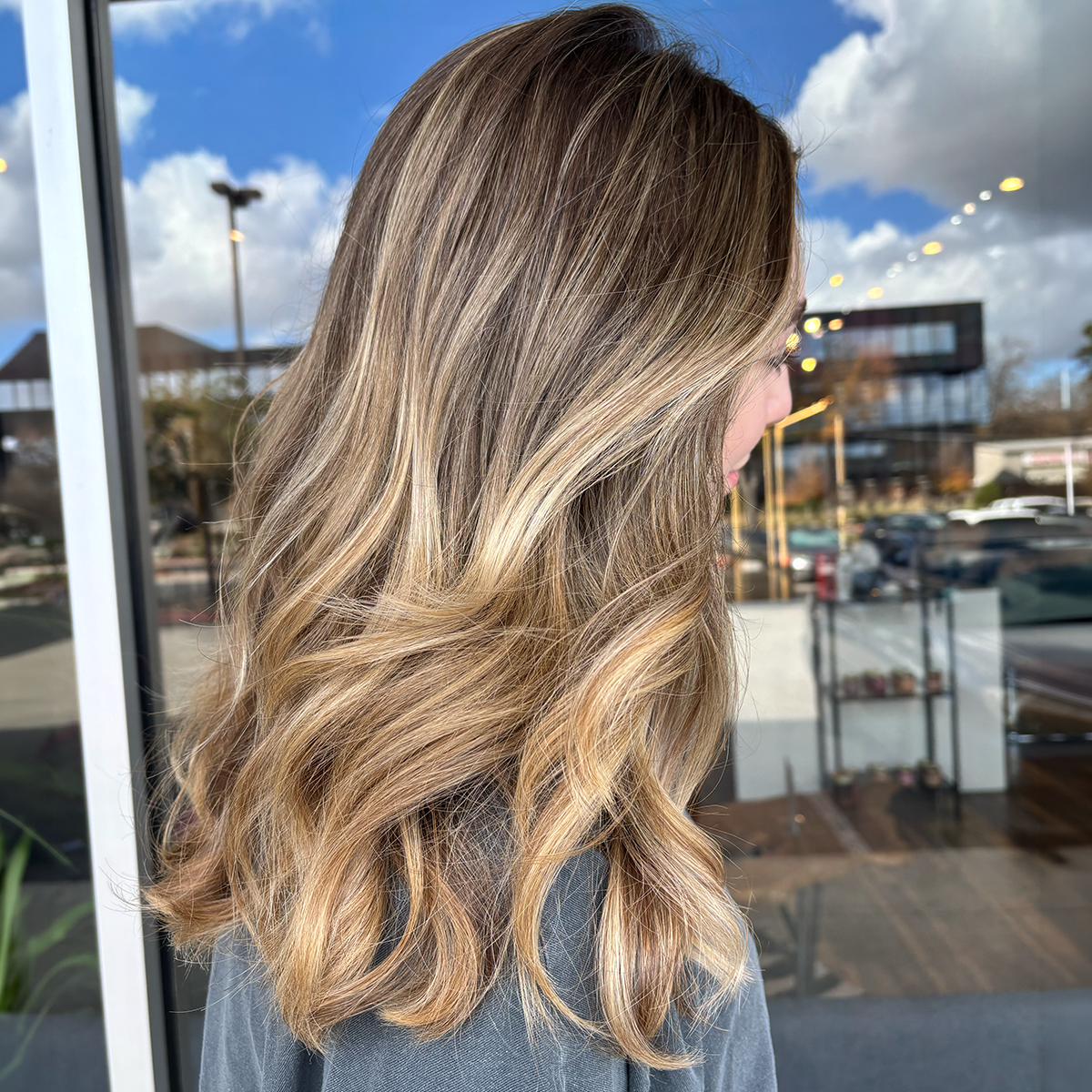 Side view of a woman with long, wavy blonde hair standing outside near a glass window, with a parking lot and modern buildings in the background.