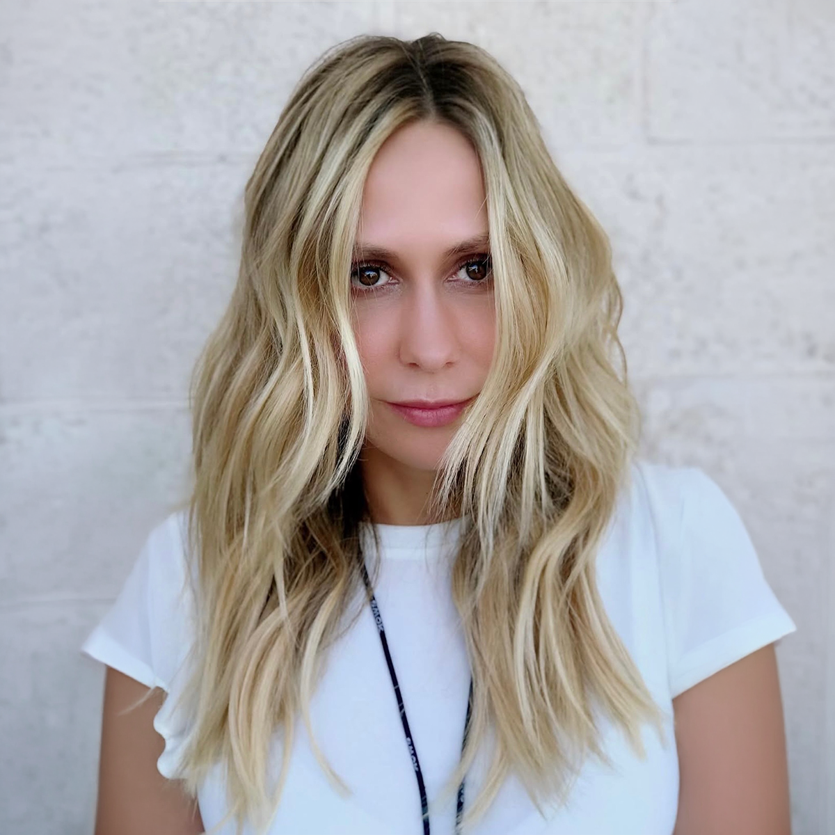 A young woman with long, wavy blonde hair and light skin, wearing a white t-shirt, standing in front of a light-colored brick wall.