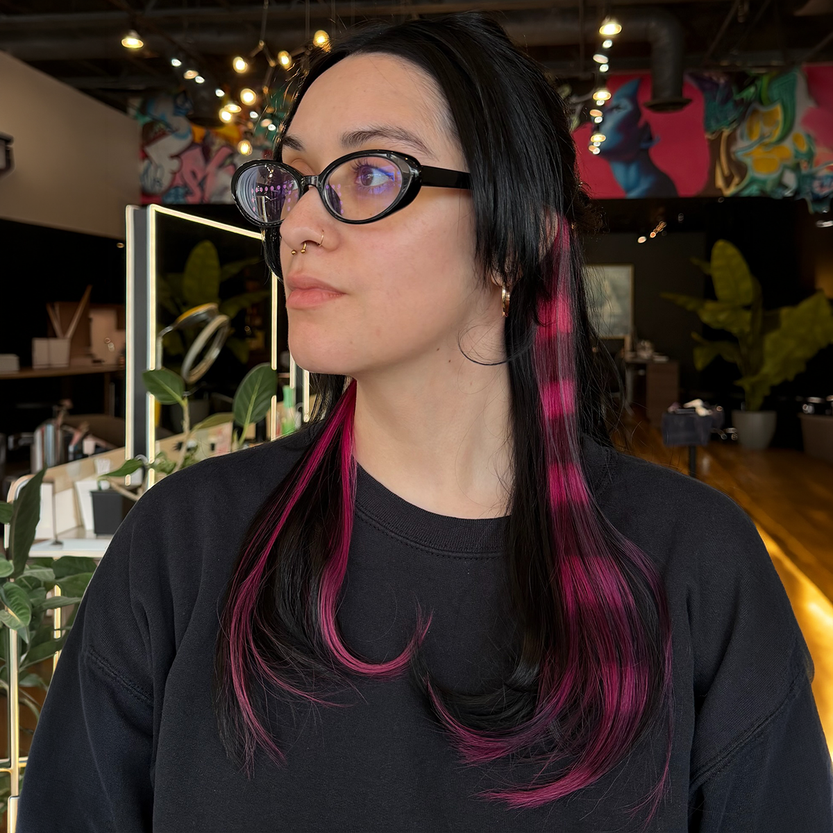 Portrait of a woman with black hair and pink highlights, wearing glasses, septum piercing, hoop earrings, and a black shirt in an indoor space with plants and colorful artwork.