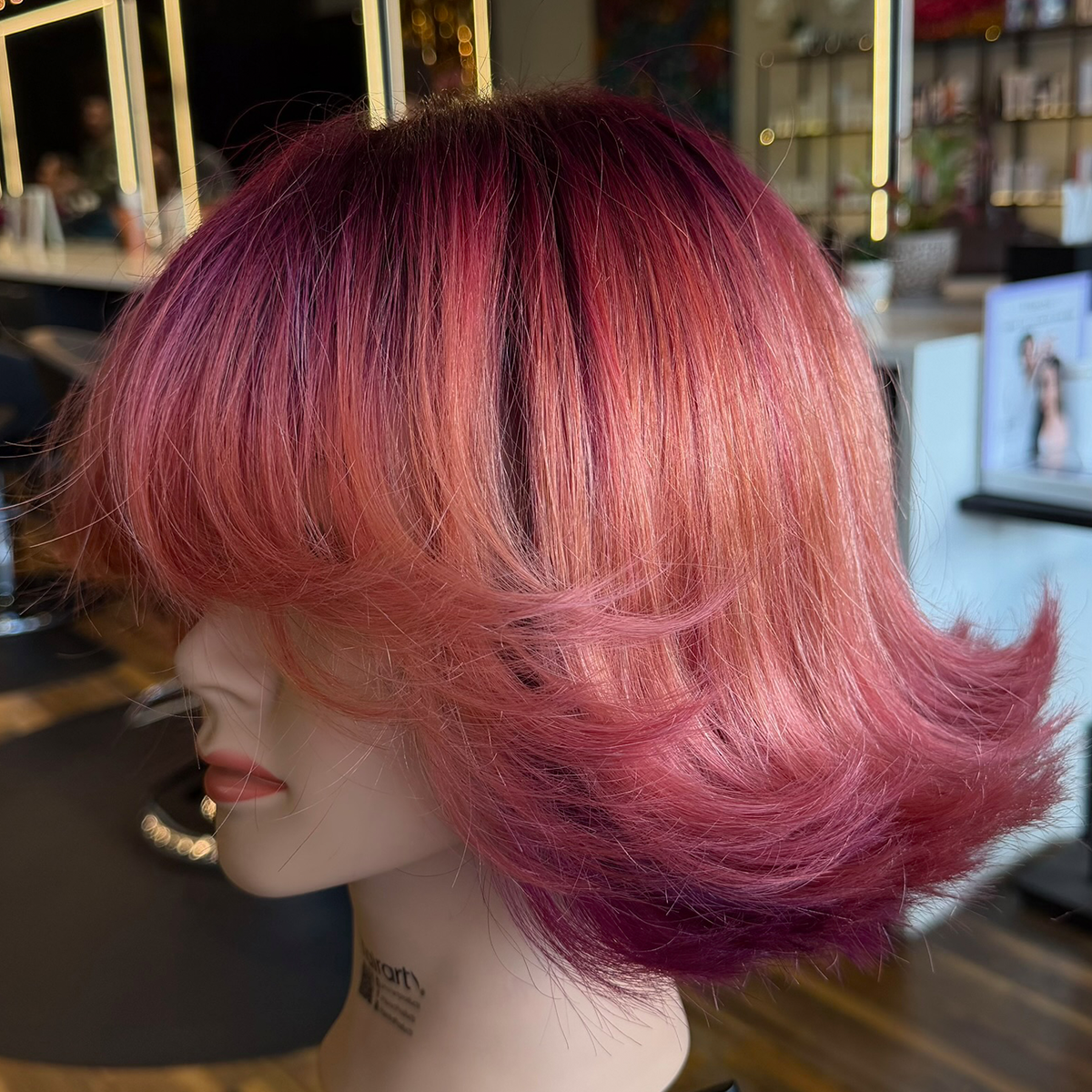 Close-up of a mannequin with a short, layered hairstyle colored in shades of pink, orange, and purple in a salon setting.