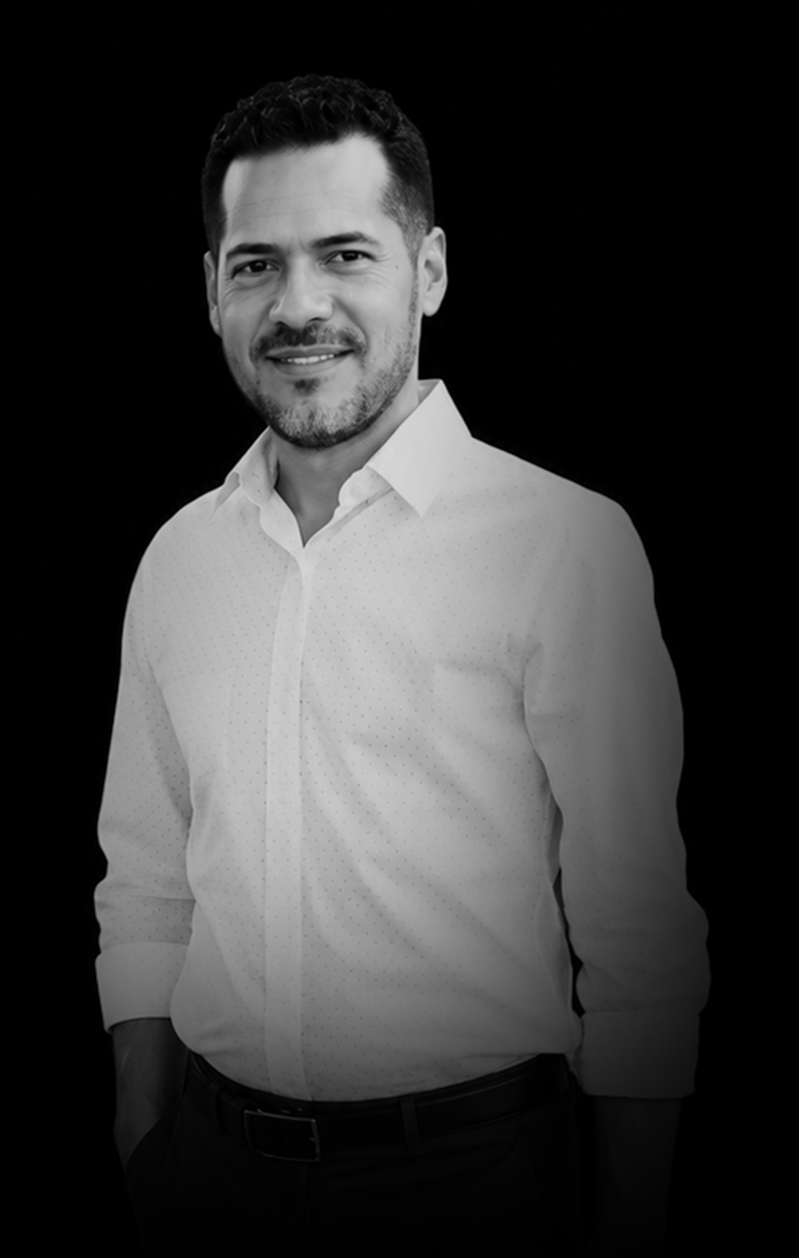 Black and white portrait of a man with short dark hair, beard, wearing a white button-up shirt, smiling, against a solid dark background.