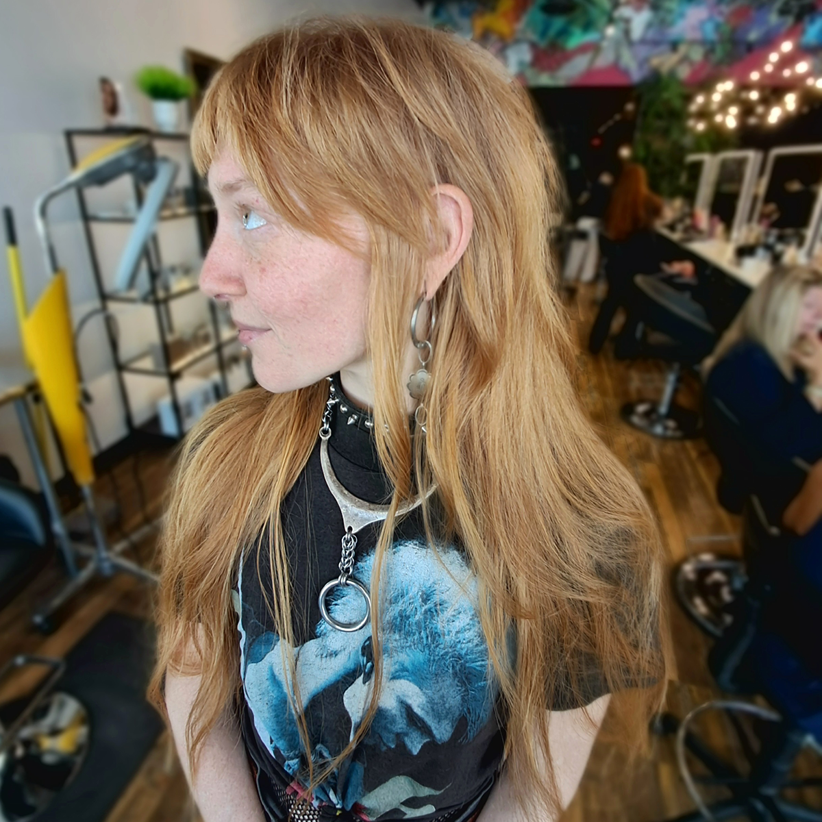 A woman with long red hair, wearing earrings and a necklace, looking to her left in a hair salon with other clients and salon equipment in the background.