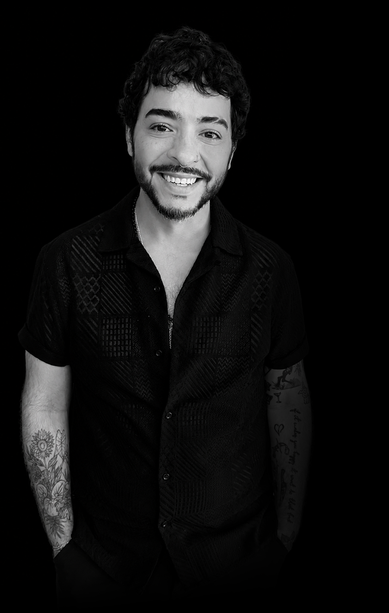 Black and white portrait of a smiling young man with curly hair, beard, and tattoos wearing a short-sleeve button-up shirt, against a dark background.