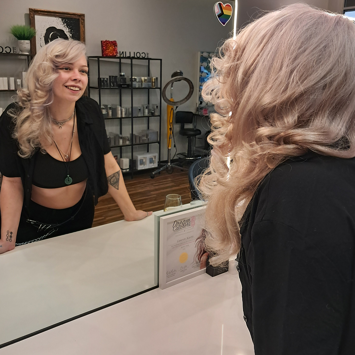 A woman with long, curly blonde hair and a black outfit is smiling at a counter, talking to another woman with similar hair style and color. The setting appears to be a salon or beauty studio with shelves of supplies in the background.