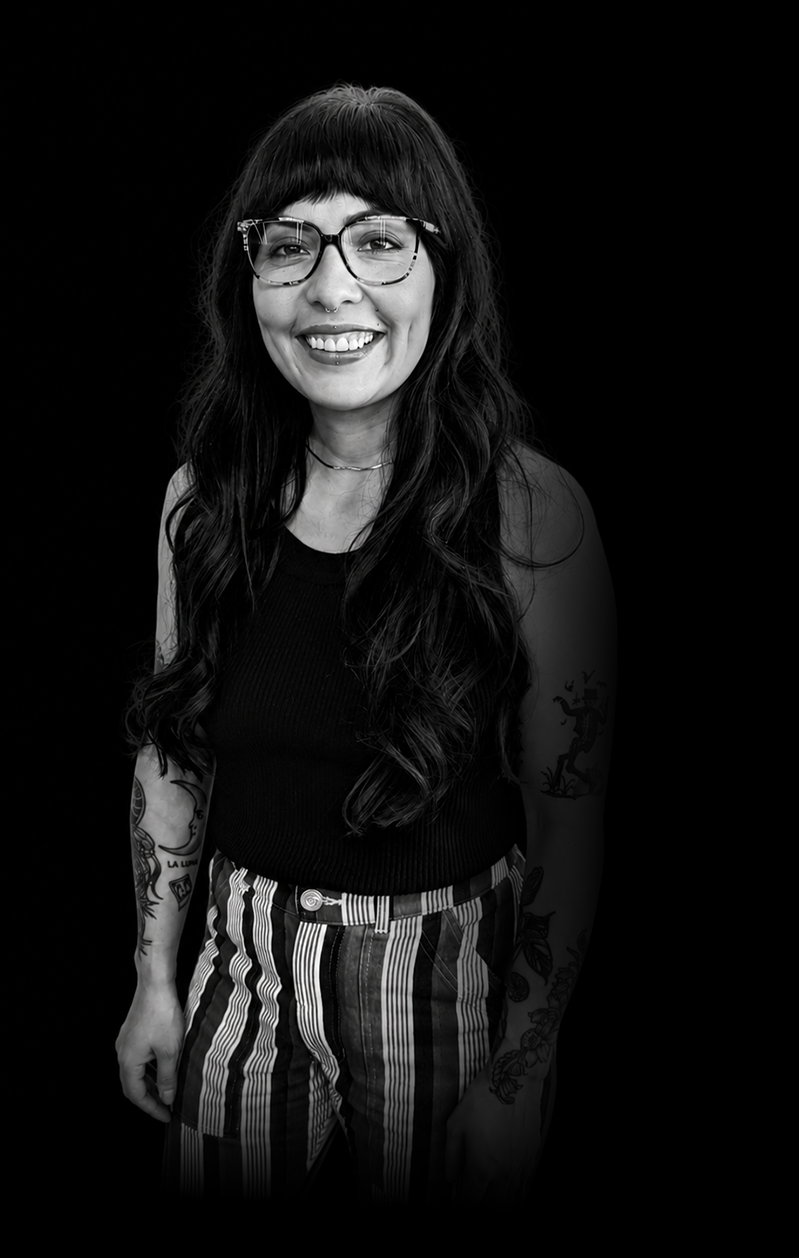 Black and white portrait of a smiling woman with long wavy hair, wearing glasses, a sleeveless top, and striped pants, standing against a dark background.