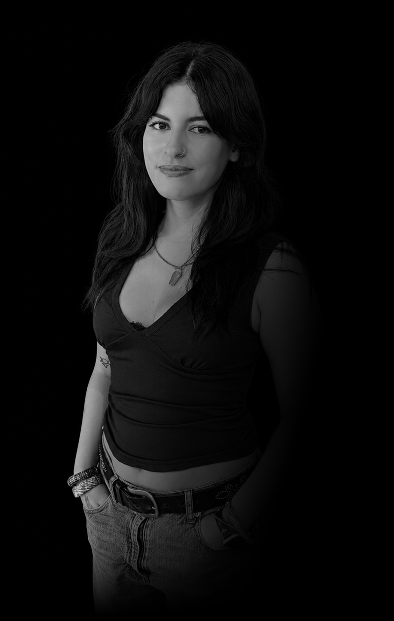 Black and white portrait of a young woman with long dark hair, wearing a sleeveless top, jeans, a belt, and jewelry, standing against a dark background.