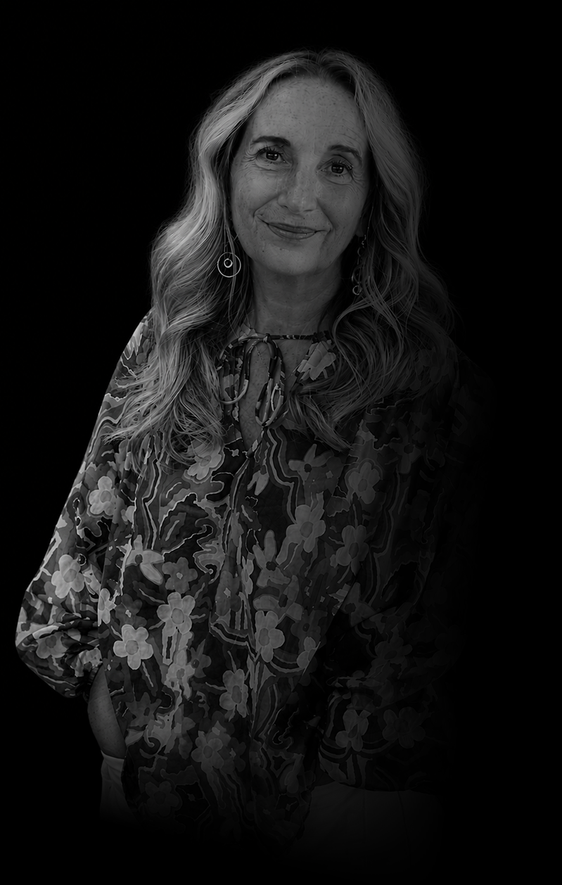 Black-and-white portrait of a woman with long, wavy hair, smiling gently, wearing patterned blouse and hoop earrings, posed against a dark background.