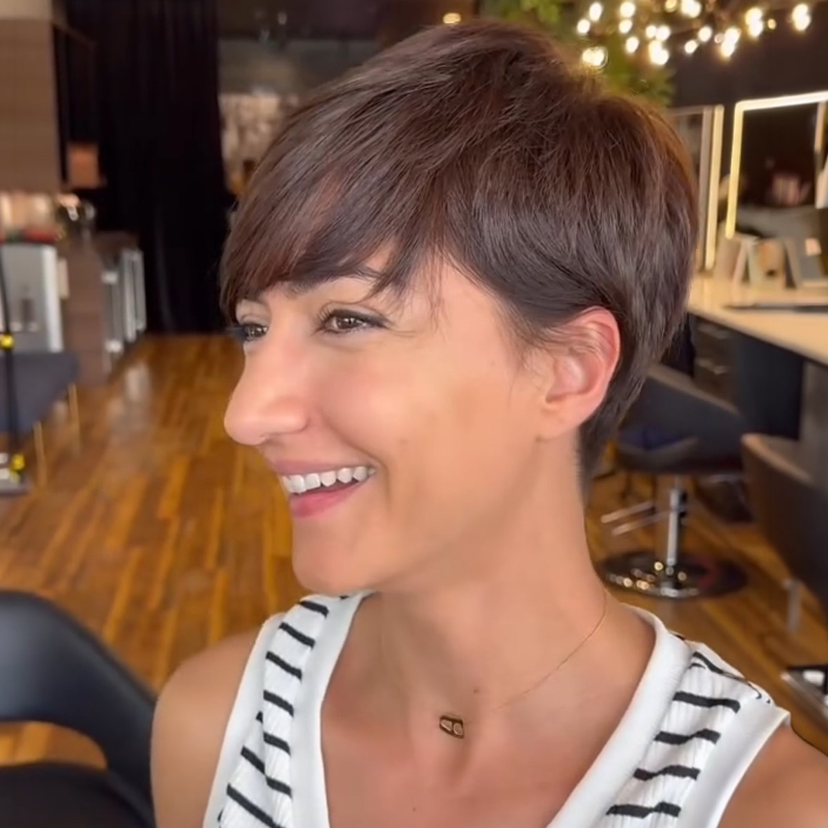 A smiling woman with short brown hair in a striped tank top in a salon or hair studio.