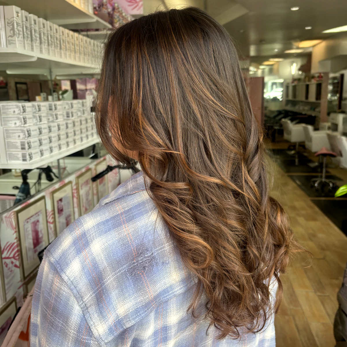 A woman with long, wavy, brown hair with caramel highlights, wearing a plaid shirt, standing in a retail store aisle.