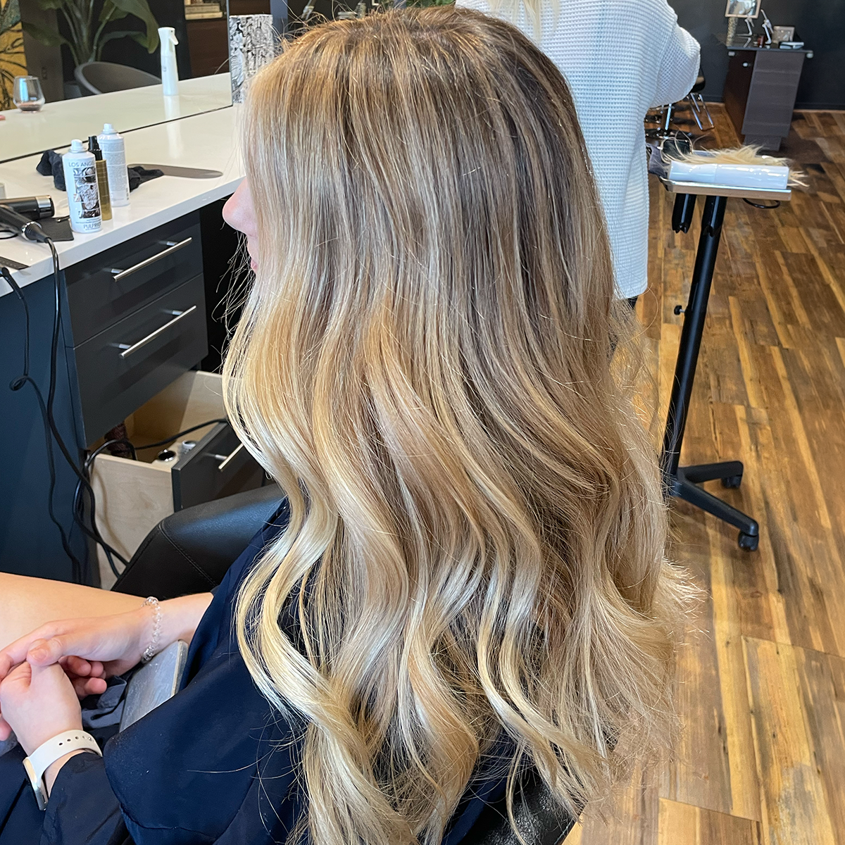 A woman with long, wavy blonde hair sitting in a hair salon chair, awaiting a haircut or styling.
