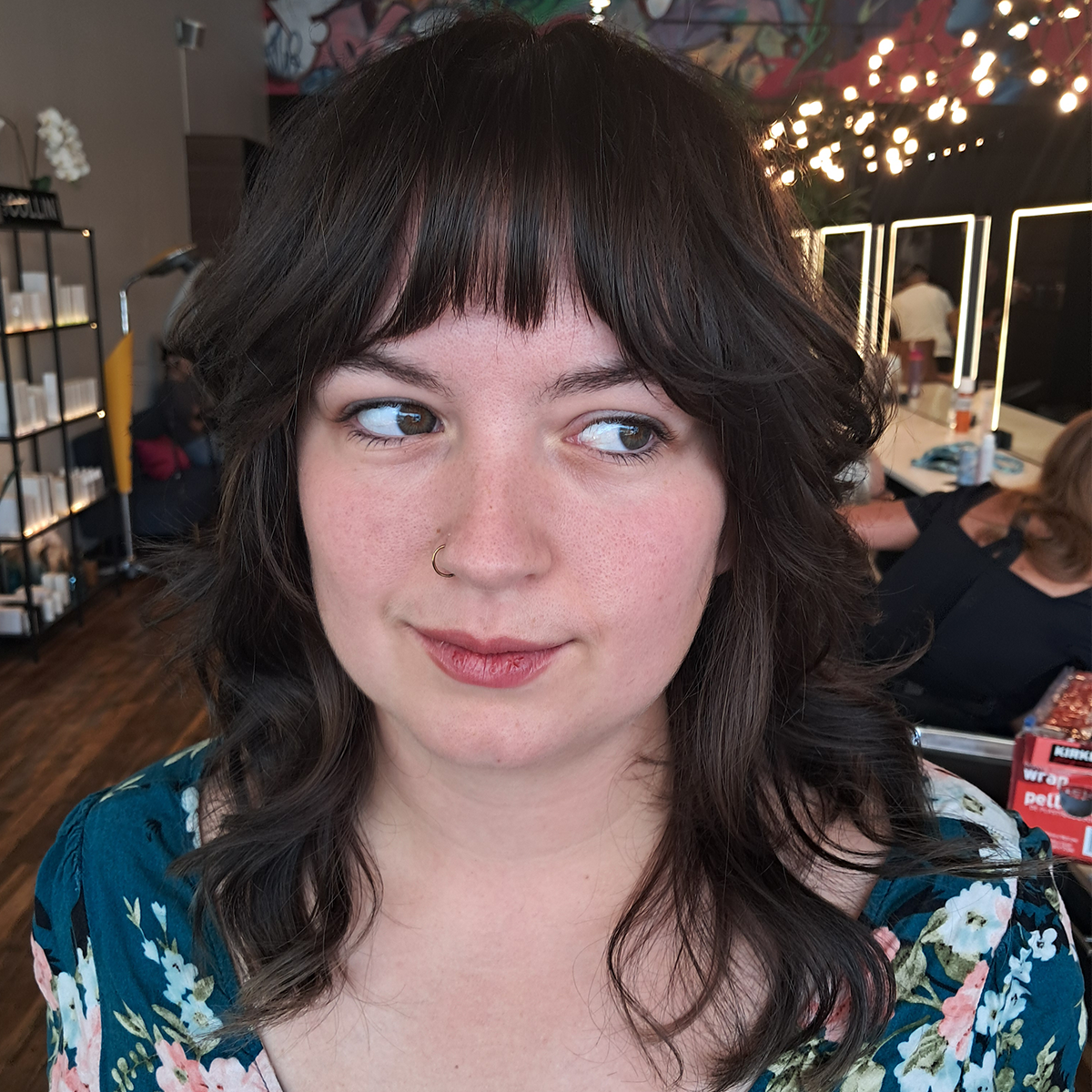 A young woman with dark curly hair and a nose ring looking to the side in a cafe, with lights and people in the background.