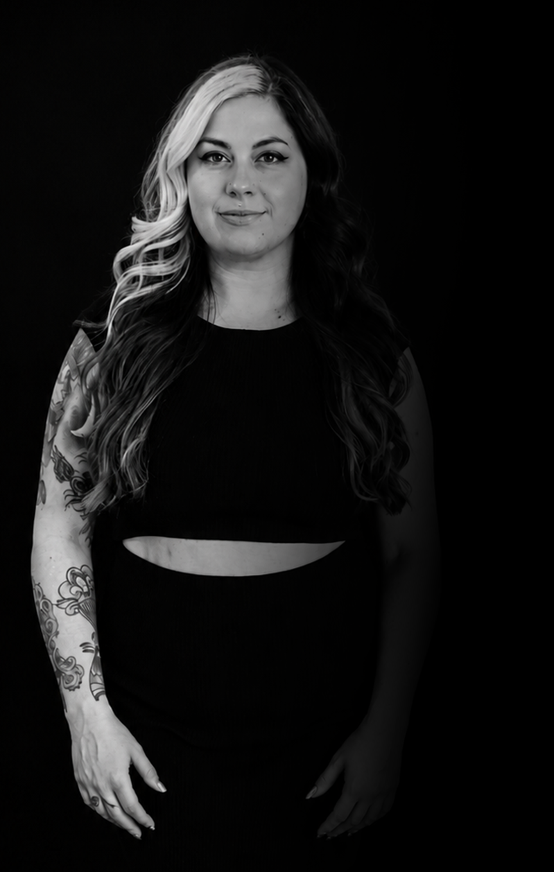 Black and white portrait of a woman with long wavy hair, wearing a sleeveless black dress with a midriff cutout, standing against a dark background.