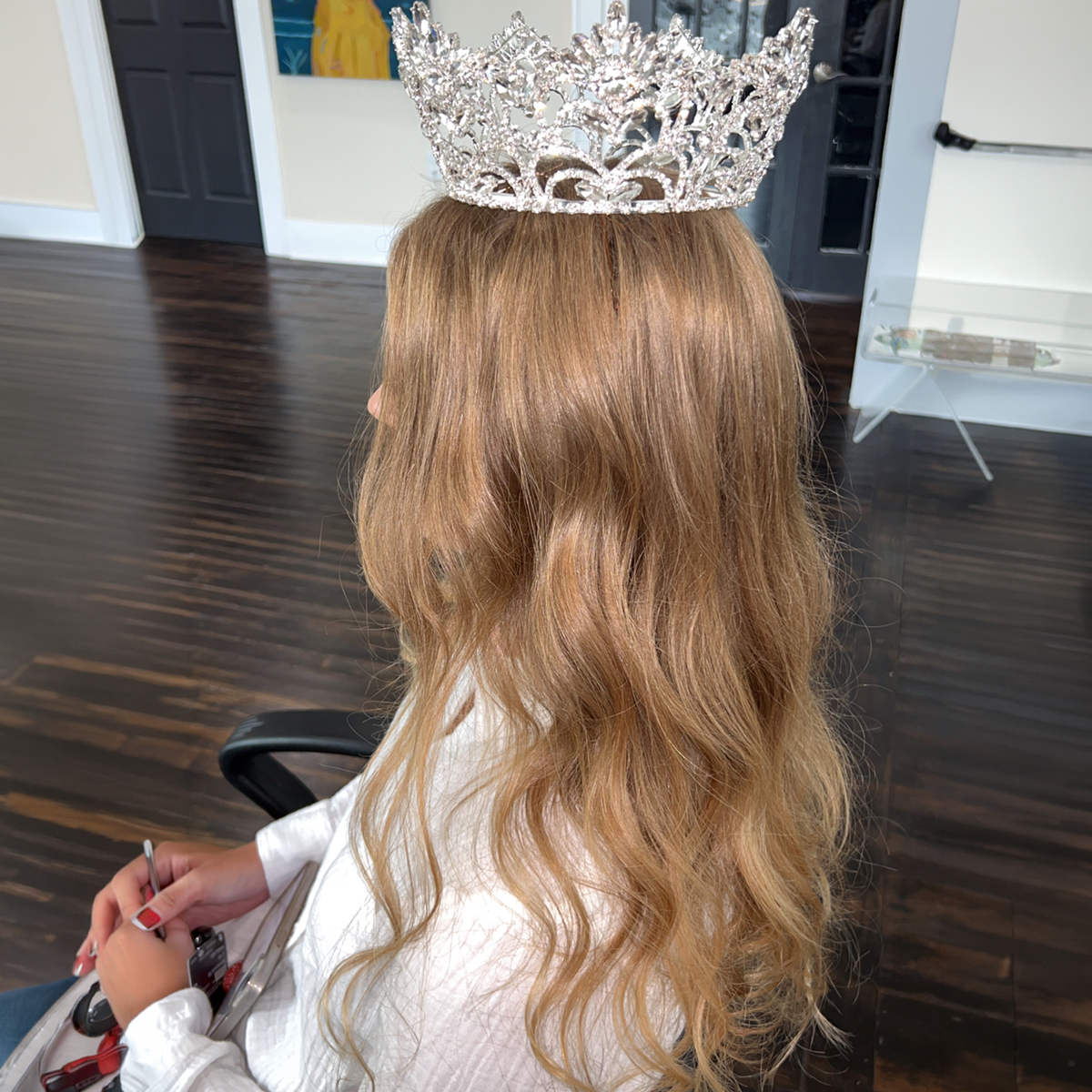 Person with long wavy blonde hair wearing a silver tiara, sitting in a chair in an indoor space with dark hardwood floors.