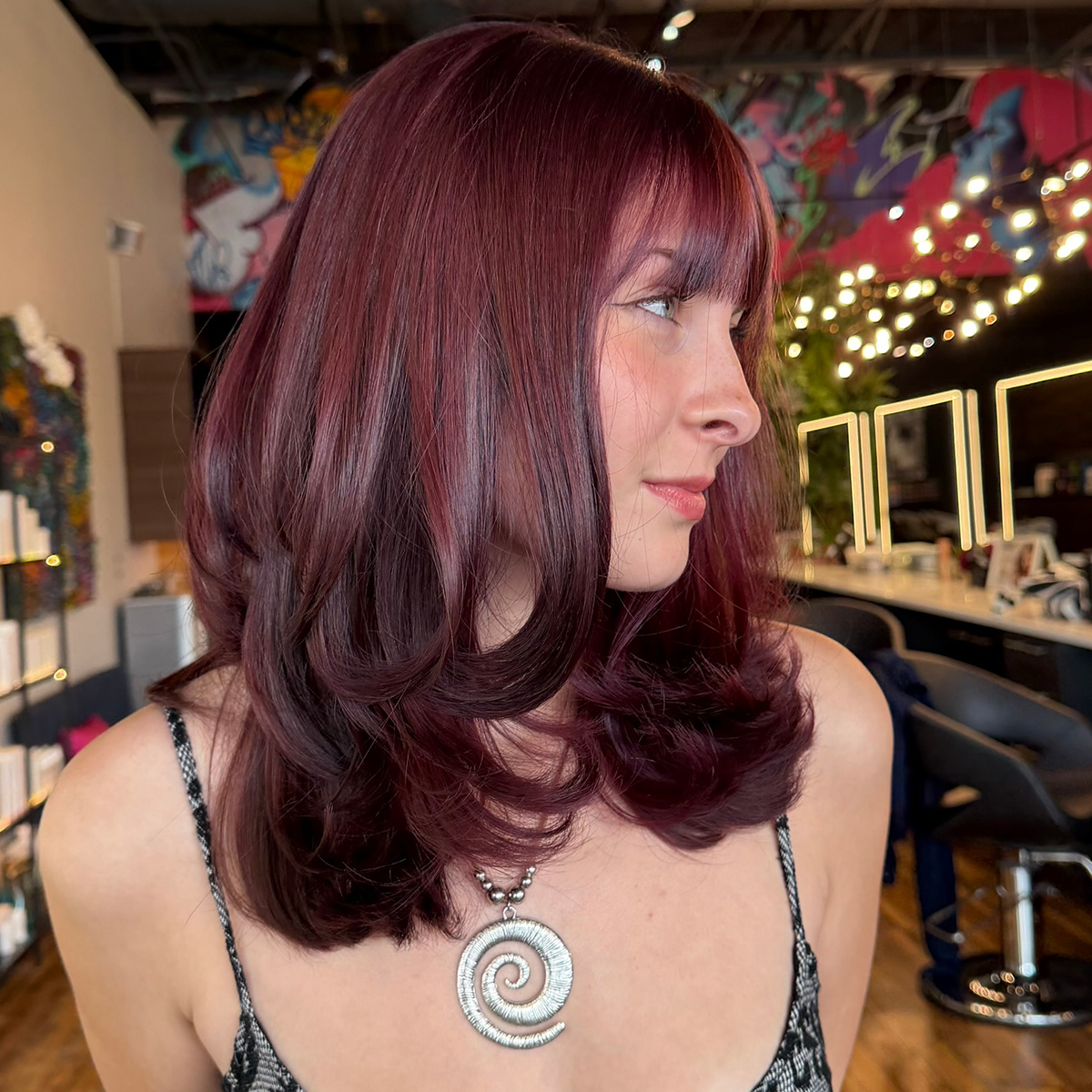 A woman with shoulder-length, wavy, reddish-brown hair wearing a spiral shell necklace in a salon or studio with colorful artwork and string lights in the background.