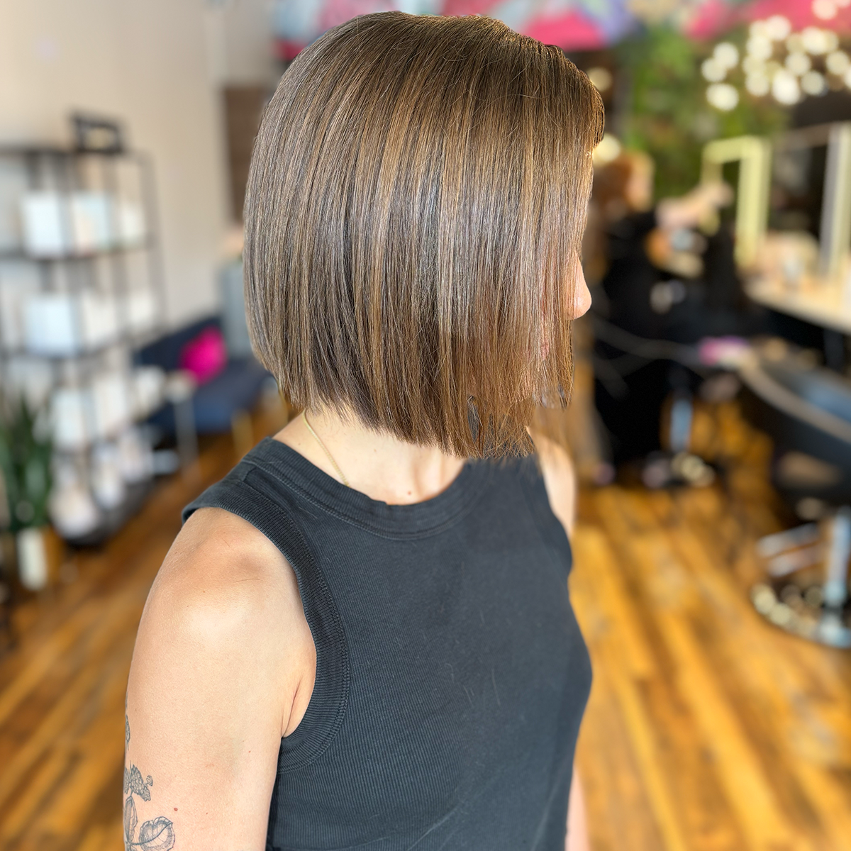 Woman with a blunt bob haircut sitting in a salon, wearing a sleeveless black top, with a blurred background of salon chairs and other clients.