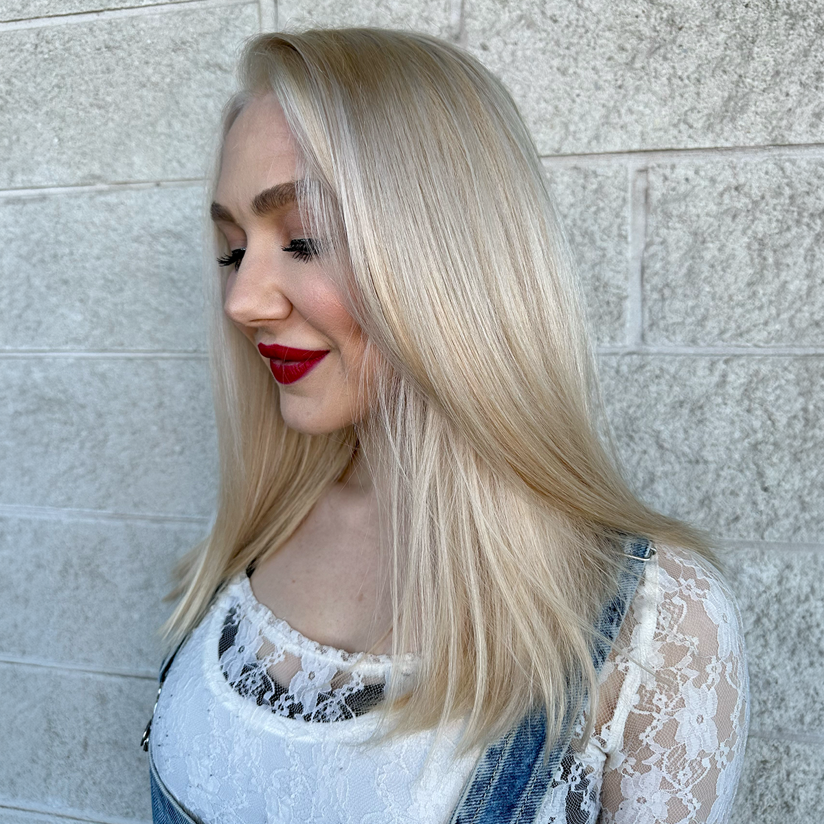A young woman with long blonde hair, red lipstick, and a lace top, looking down and smiling, standing against a light-colored brick wall.