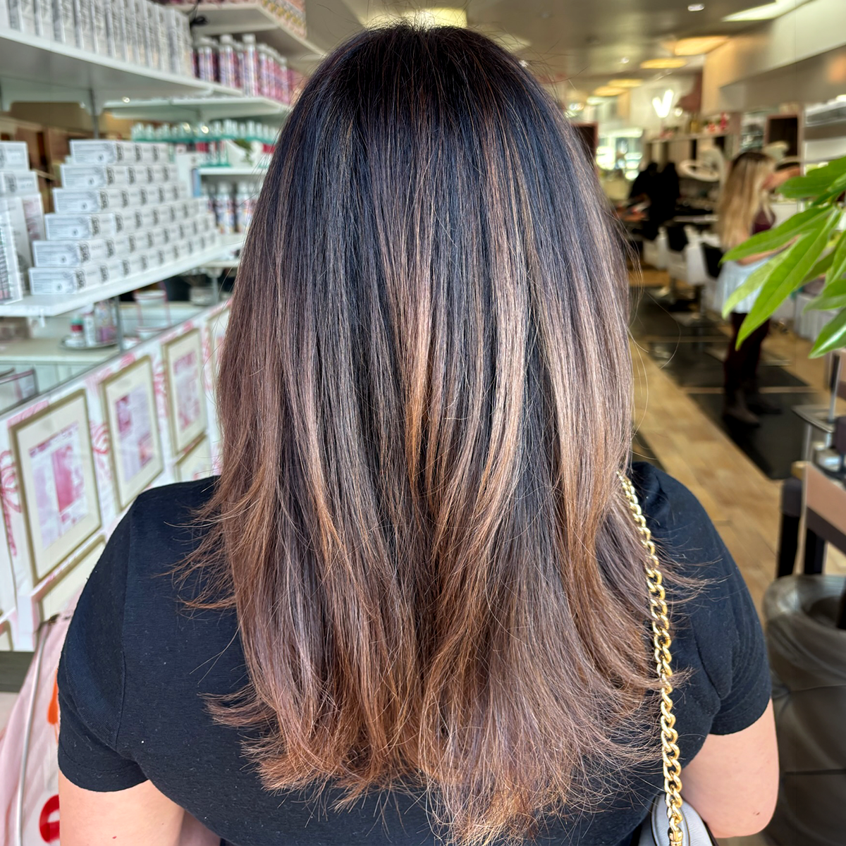 Back view of a woman with shoulder-length brown hair with blonde highlights, wearing a black top and a gold chain strap purse, in a store or salon with shelves and other people in the background.