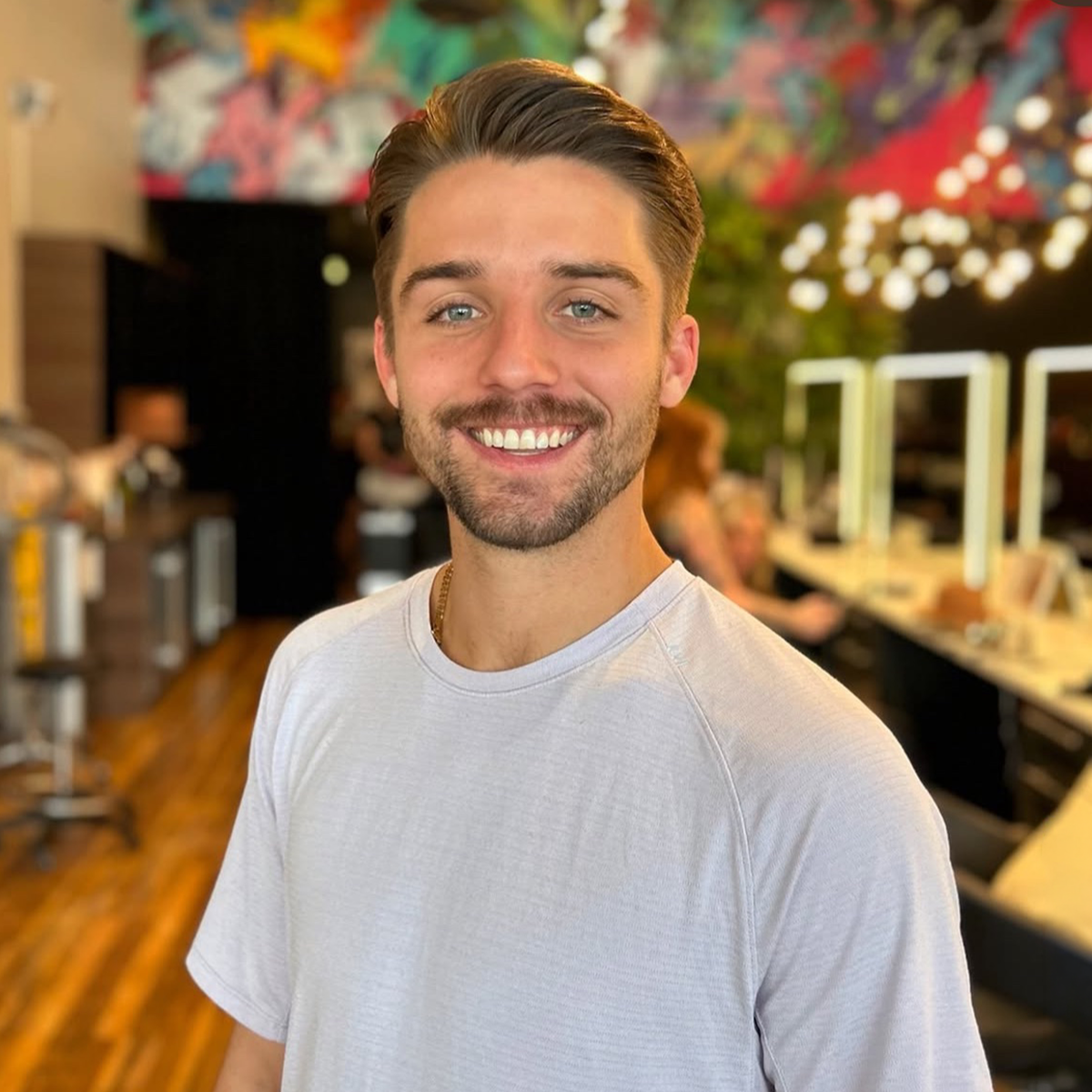 Smiling young man with short dark hair and a beard, wearing a white t-shirt, standing in a vibrant interior space, possibly a cafe or a workspace, with people working at tables and colorful wall art in the background.