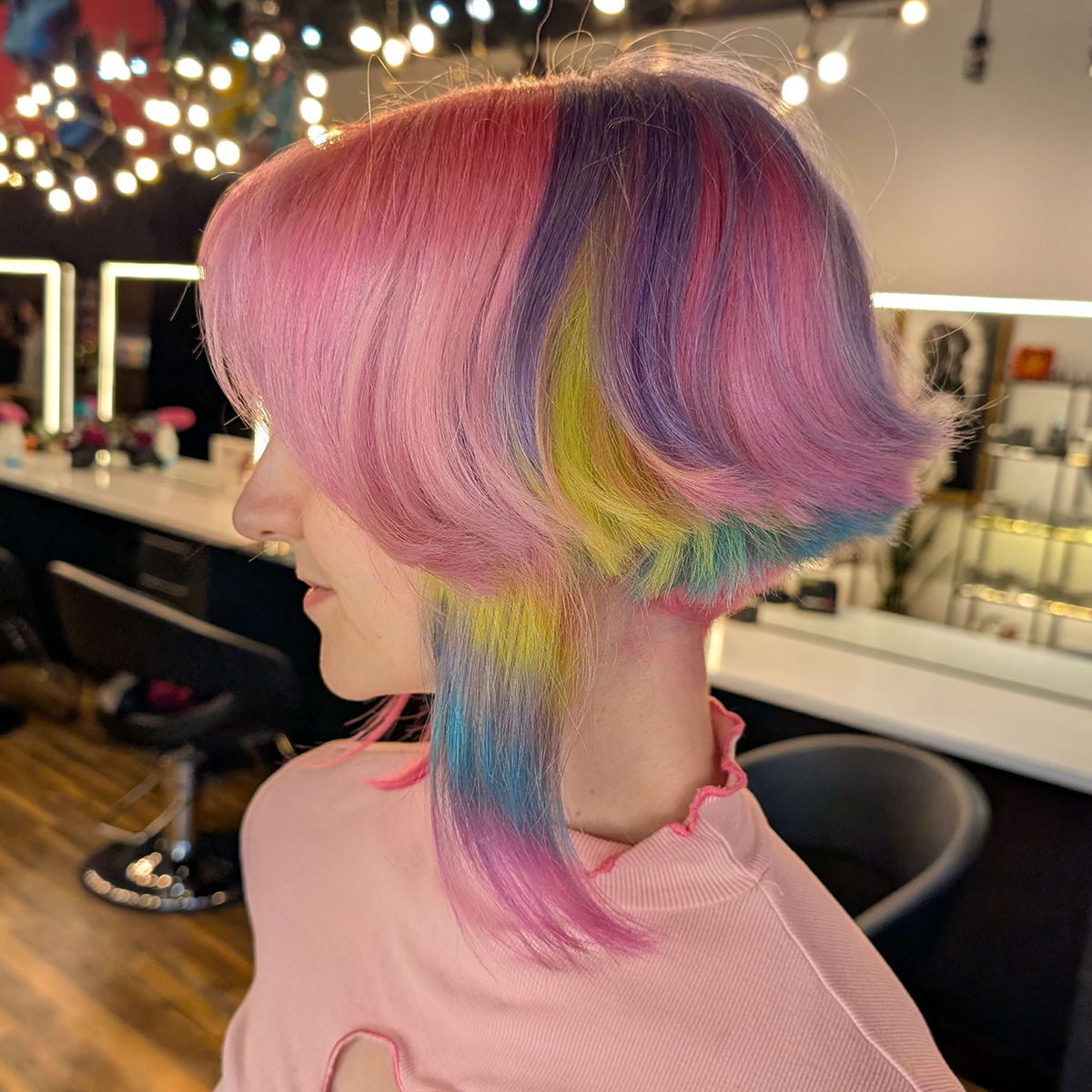Side view of a woman with a colorful, multi-hued bob hairstyle featuring pink, purple, yellow, green, and blue shades, inside a hair salon with illuminated mirrors and salon equipment in the background.
