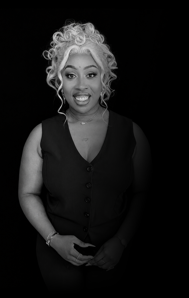 A woman with curly blonde hair, wearing a sleeveless buttoned vest, smiling and posing against a black background.