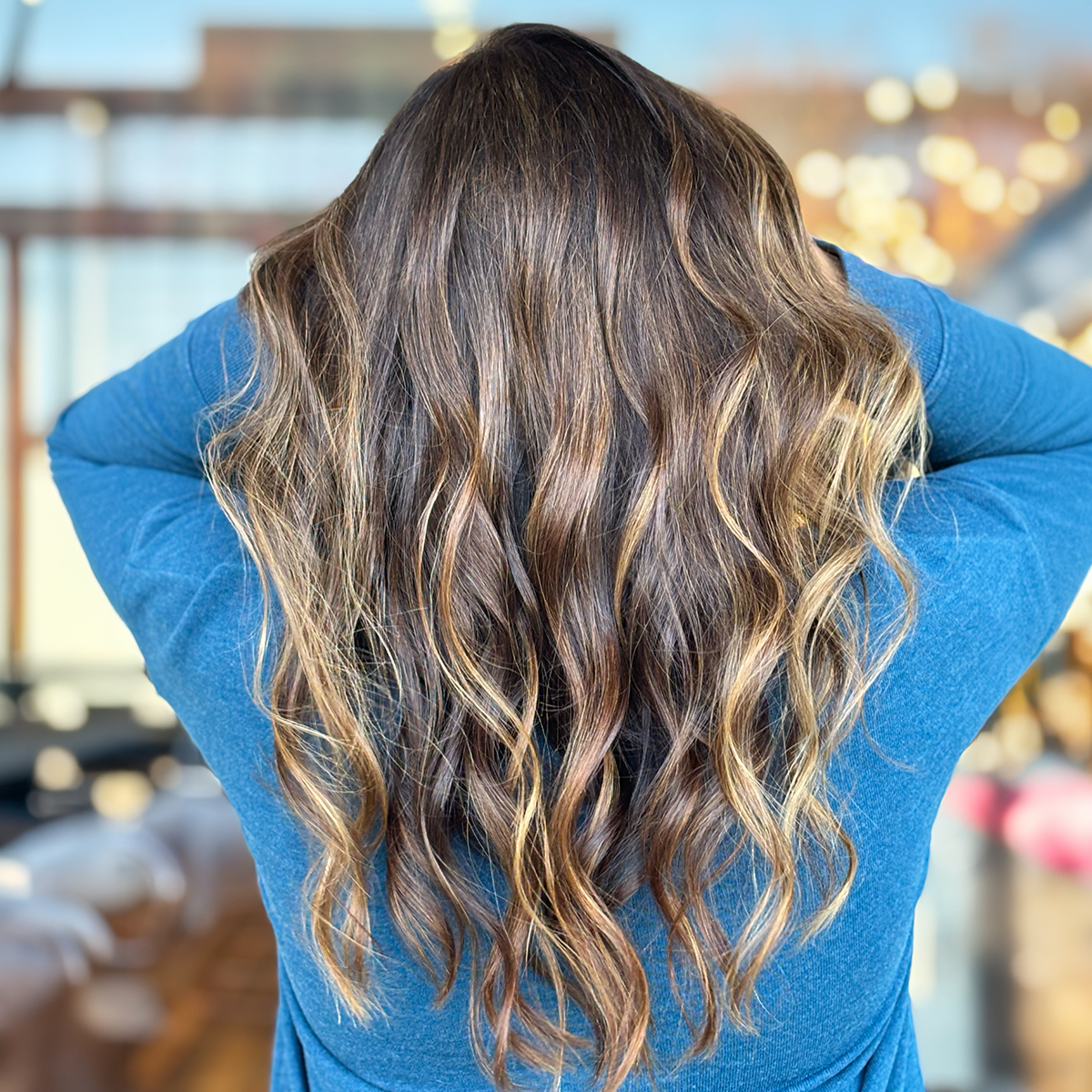 Person with long wavy hair, seen from behind, wearing a blue shirt, in an indoor setting with blurred warm lighting in the background.