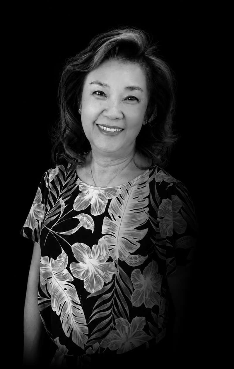 Black and white portrait of a woman smiling, wearing a tropical-patterned shirt, with wavy hair, set against a dark background.