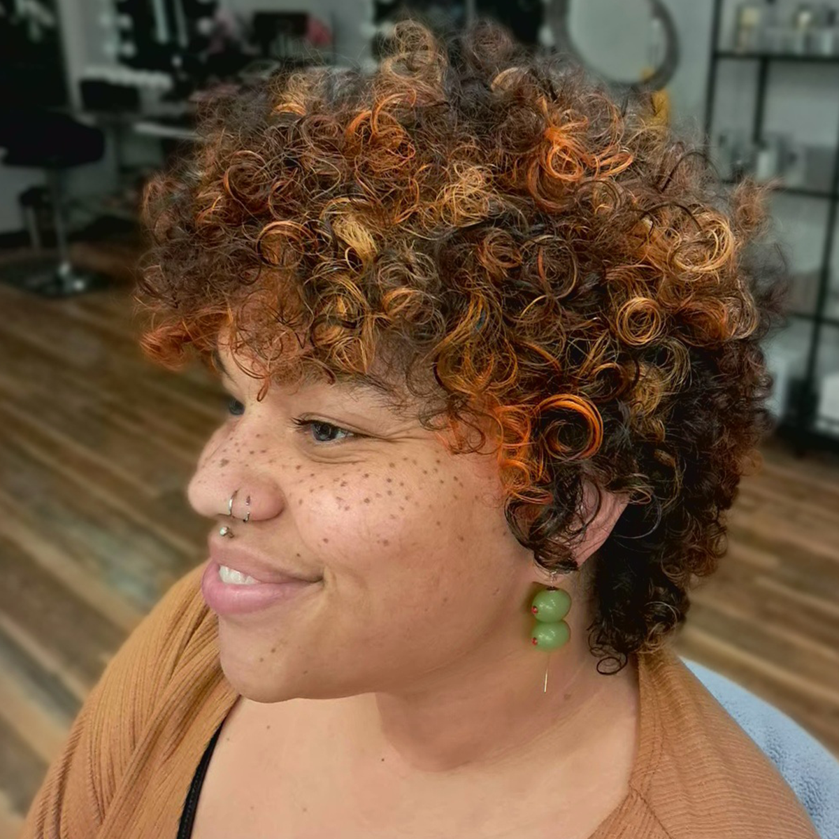 A woman with short, curly, multi-colored hair, freckles, and wearing green earrings and a nose ring, smiling in an indoor setting.