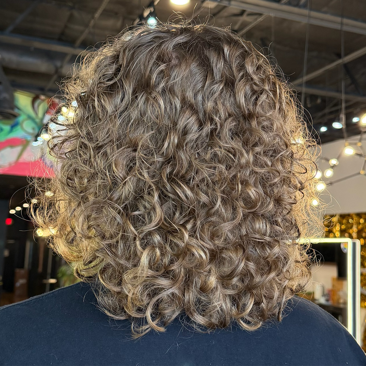 Back view of a person with shoulder-length, light brown, heavily curly hair in an indoor setting with string lights and colorful decorations.