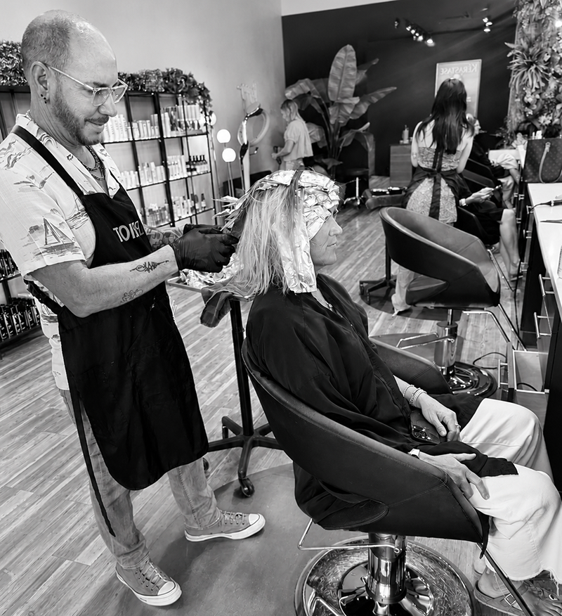 A hairdresser with tattoos and glasses applying hair dye or product to a woman's hair with foil rollers in a salon. The woman, sitting in a chair, has a salon cape on and is relaxed. In the background, other clients and hairdressers are engaged in hair treatments, with salon products and decor visible.