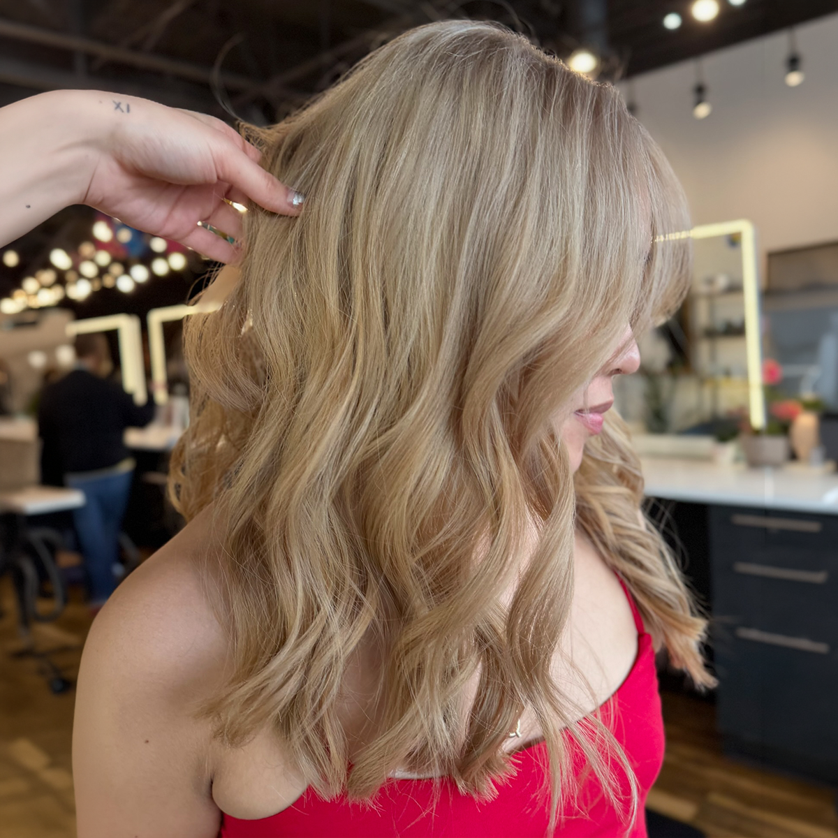 A woman with wavy blonde hair wearing a red top getting her hair styled in a salon.