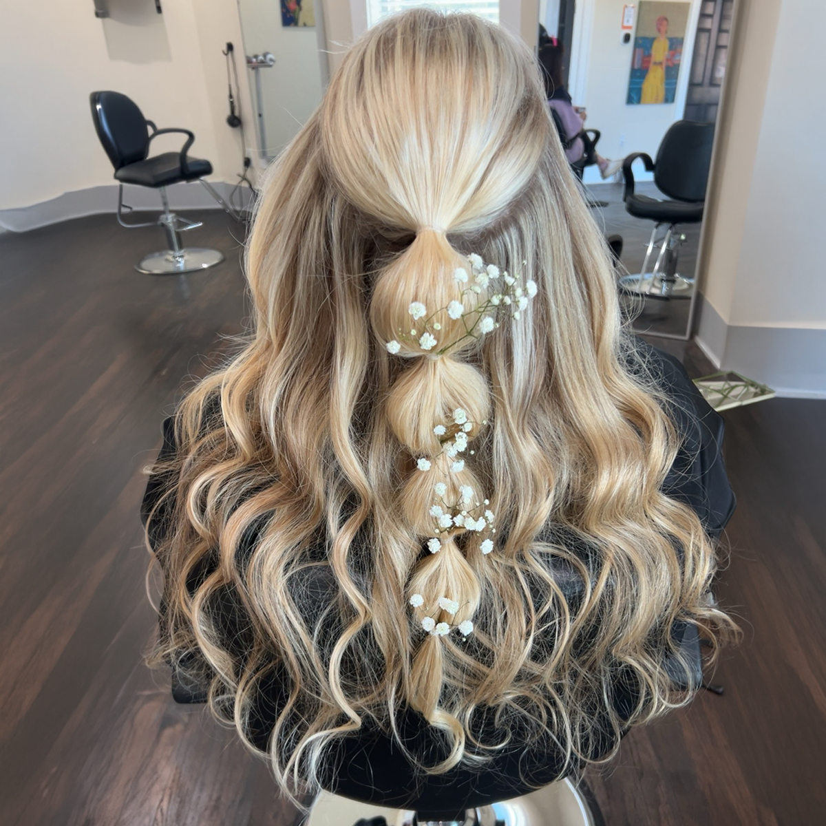 Back view of a woman with long, wavy blonde hair styled in a half-up, half-down hairstyle decorated with small white flowers.