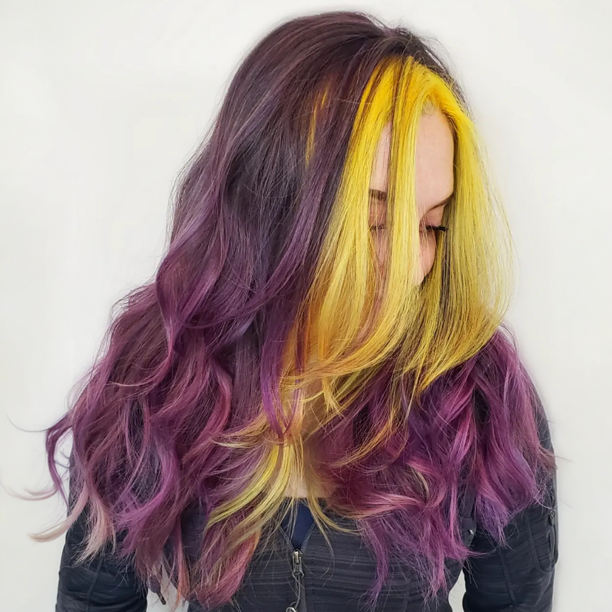 Woman with long wavy hair dyed in purple, yellow, and blonde colors, with the yellow and blonde sections framing her face and the purple on the lower ends, looking down.