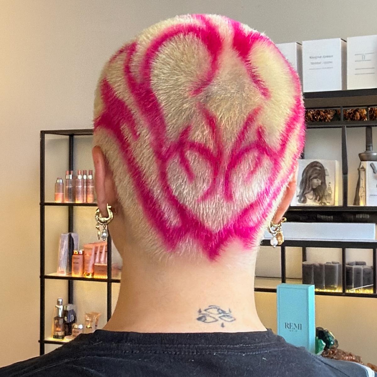 Back of a person's head with shaved hair featuring pink graffiti-style writing and design, earrings, and a tattoo on the neck, in a room with shelves holding beauty products and decor.