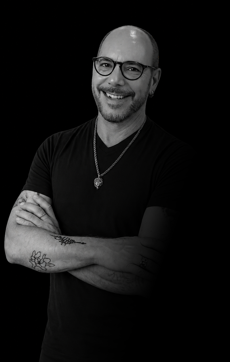 Black and white portrait of a smiling man with glasses, tattoos on his arms, wearing a black T-shirt, a necklace with a pendant, and earrings, standing against a black background.