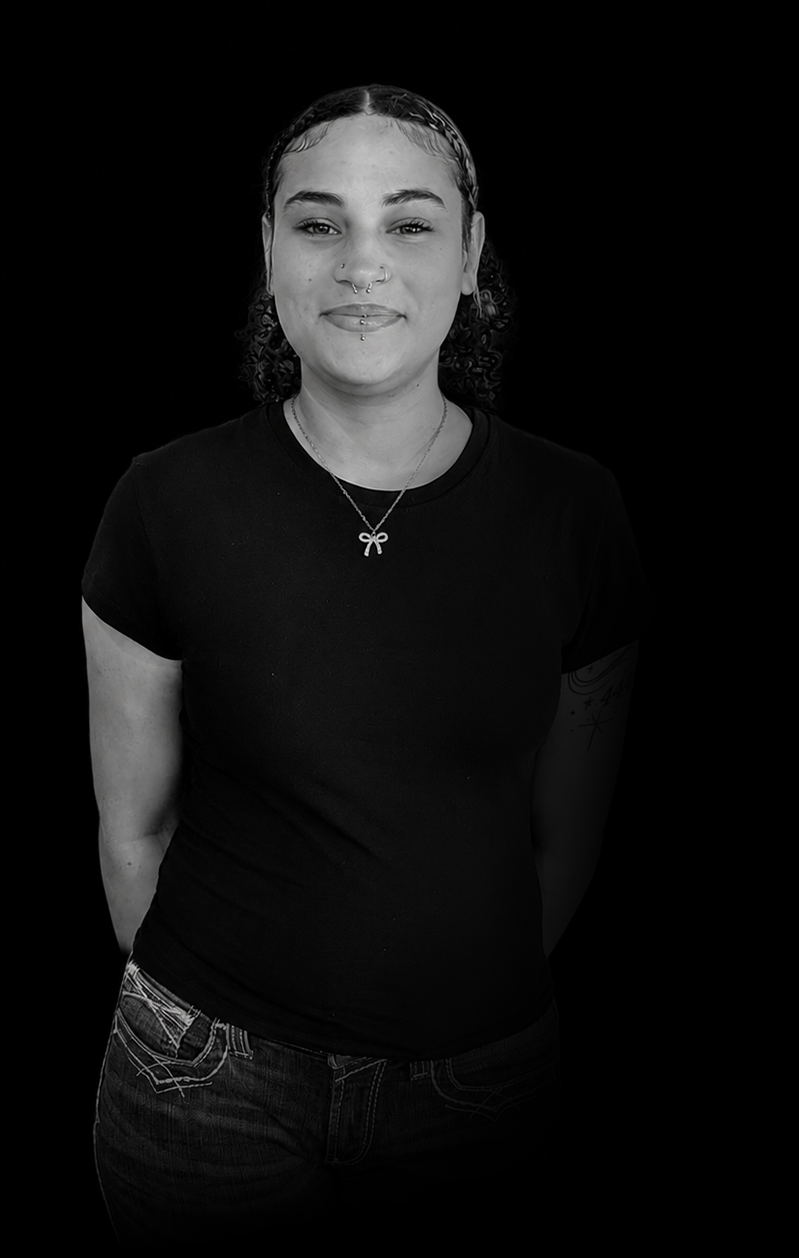 Portrait of a young woman with curly hair, wearing a black t-shirt, a necklace with a bow pendant, and earrings, standing against a black background.