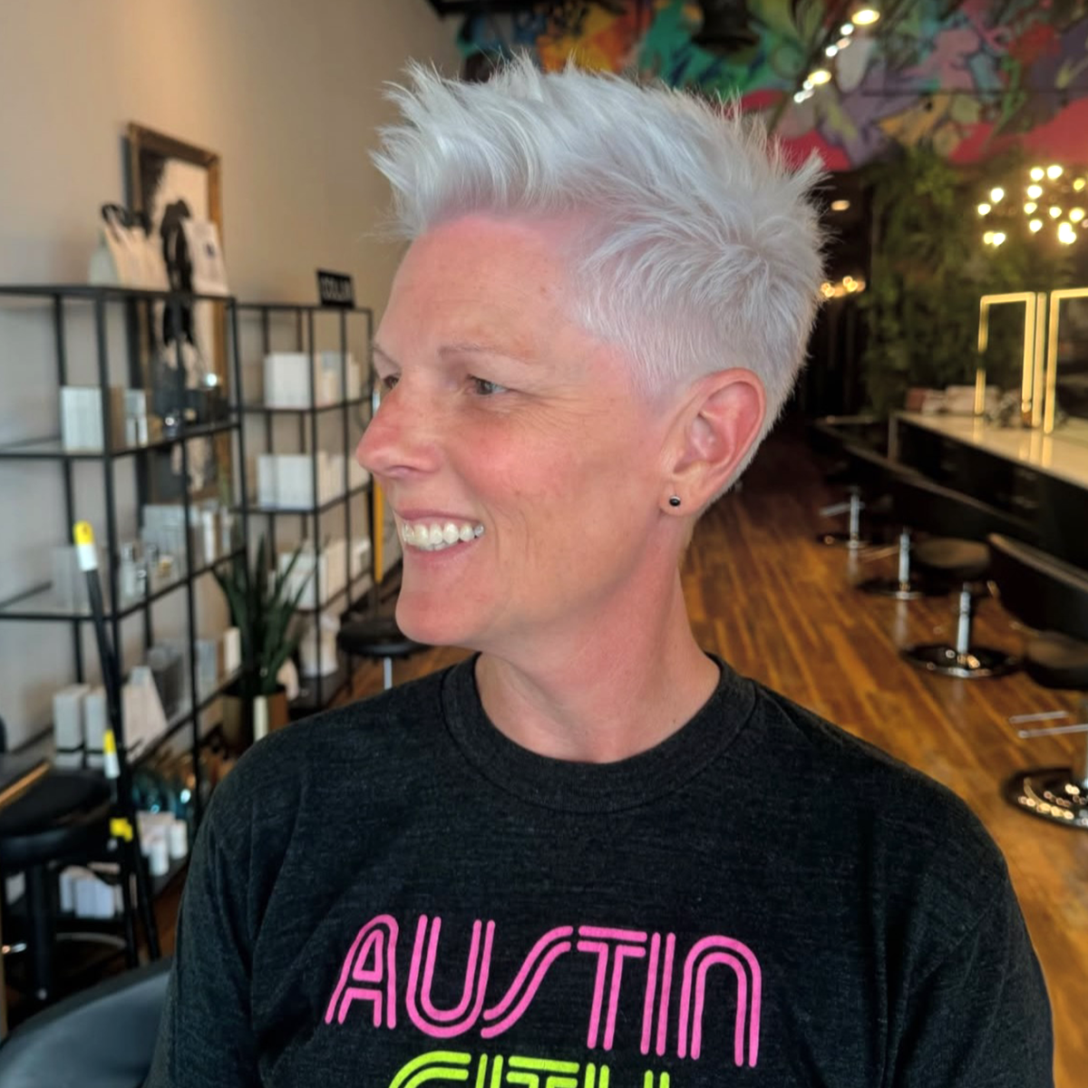A smiling woman with short platinum blonde hair styled in a spiky manner, wearing a black T-shirt with pink and green text, inside a modern hair salon with wooden floors, black chairs, and decorative shelving.