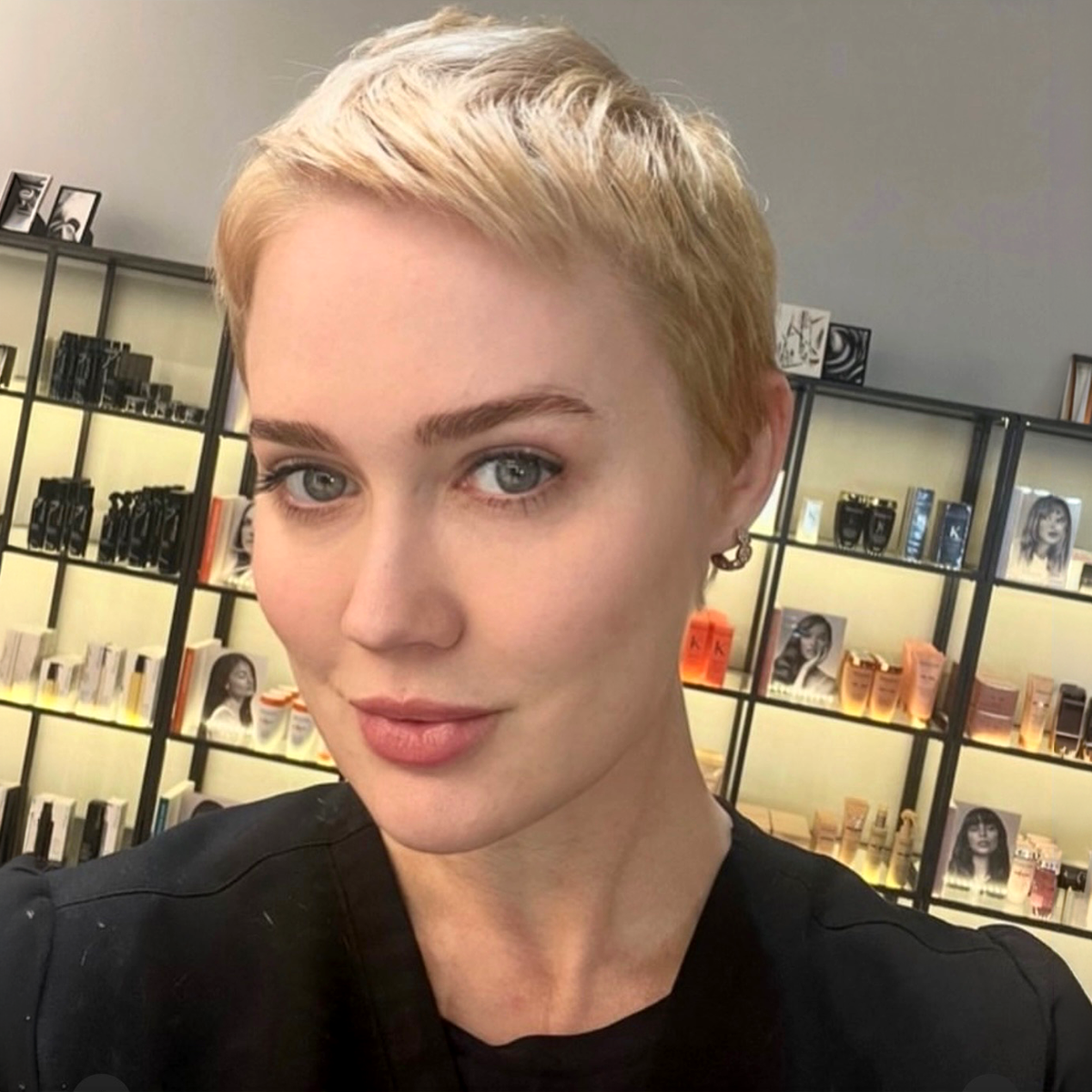 Close-up of a woman with short blonde hair and blue eyes, smiling slightly, in a store with shelves of various beauty products and books in the background.