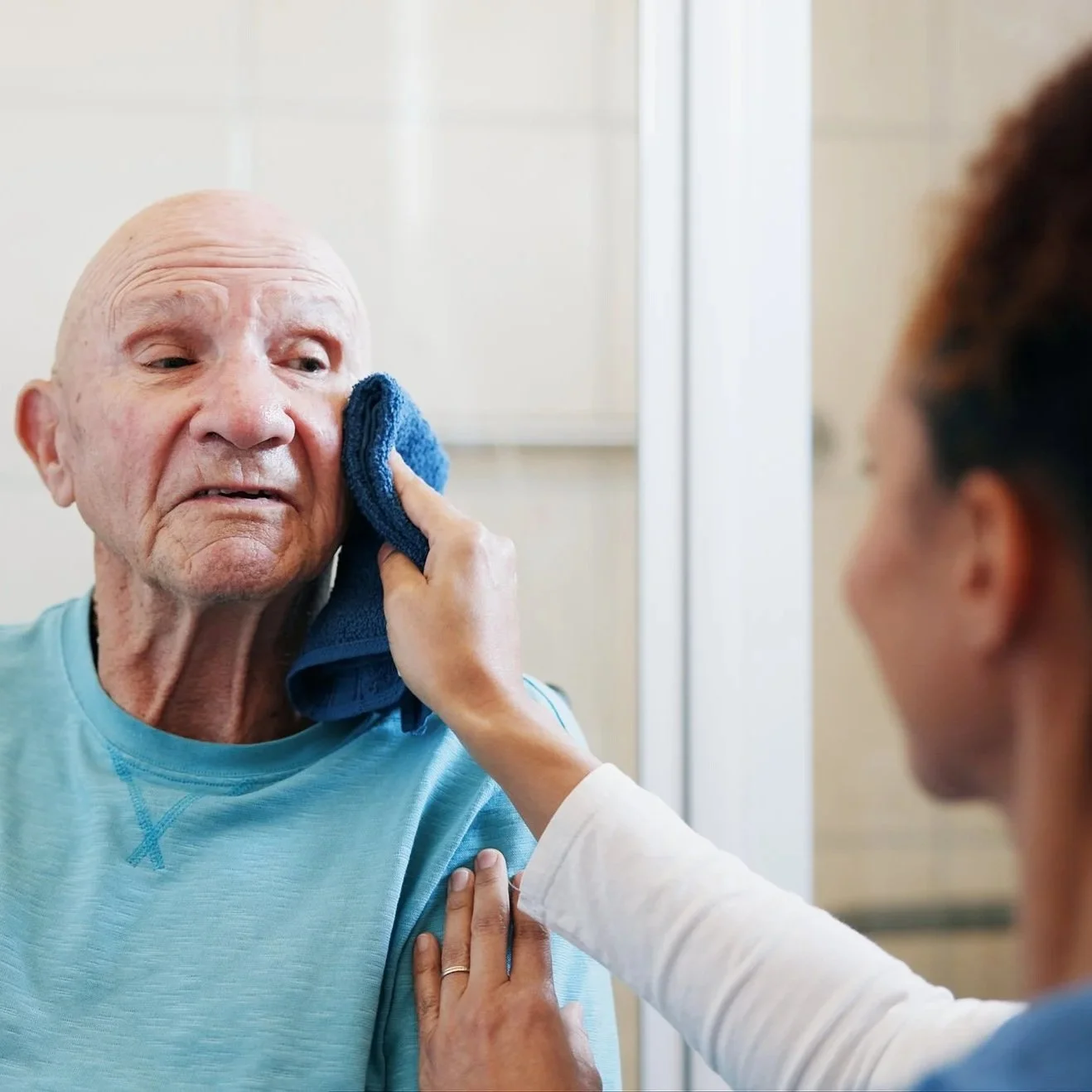 An elderly man with a wrinkled face is receiving care, gently wiping his face with a blue cloth in a healthcare setting while a caregiver supports him by holding his arm.