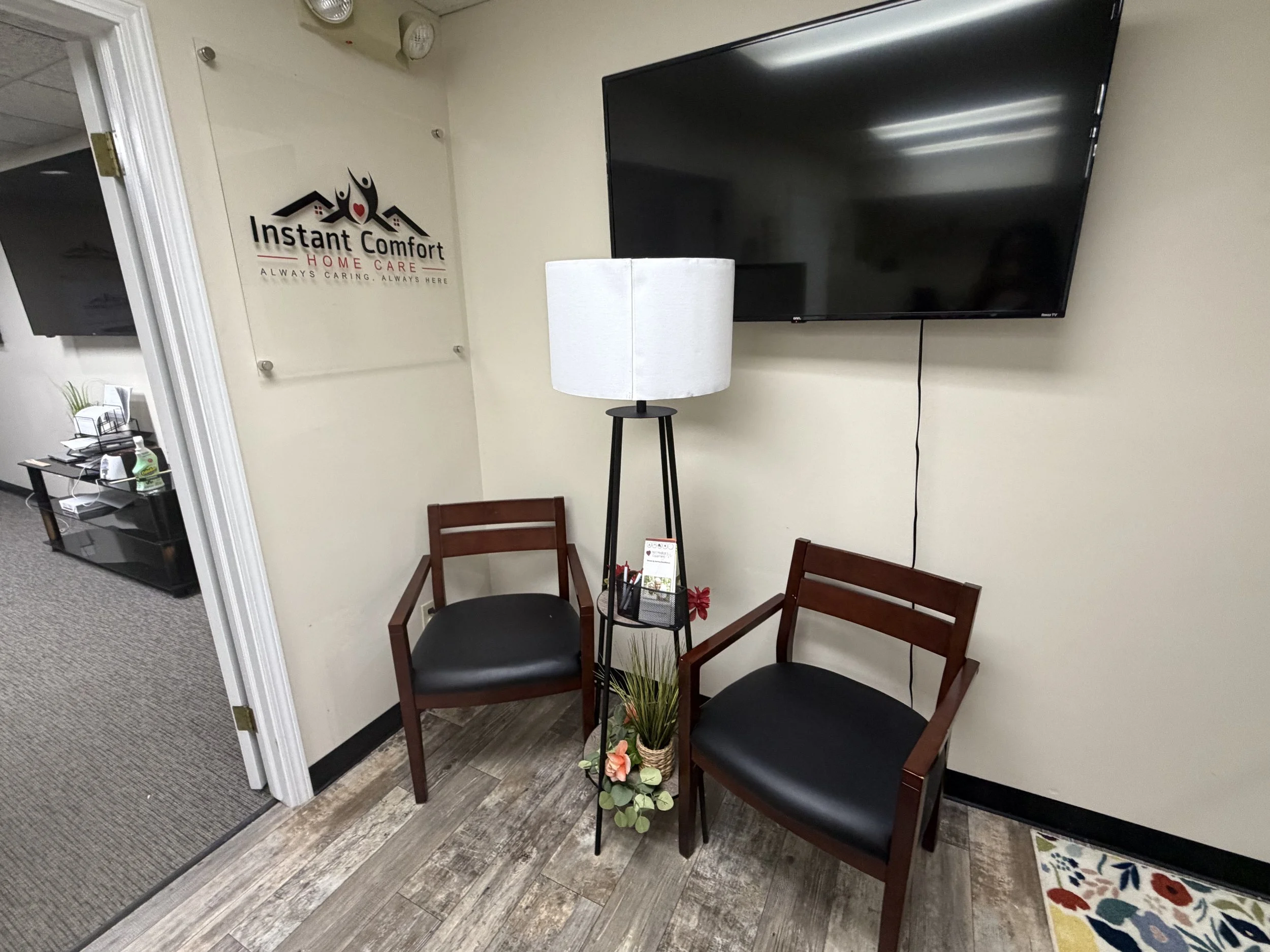 Waiting room with two black chairs and a small table with a plant and flowers. A large flat-screen TV is mounted on the wall. A sign reads 'Instant Comfort Home Care' with the slogan 'Always caring, always here'.