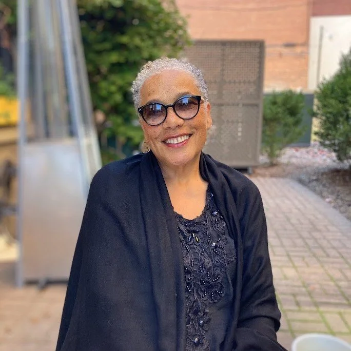 Smiling elderly woman with short curly gray hair, sunglasses, wearing a black lace top and black shawl, outdoors in a garden or patio setting.
