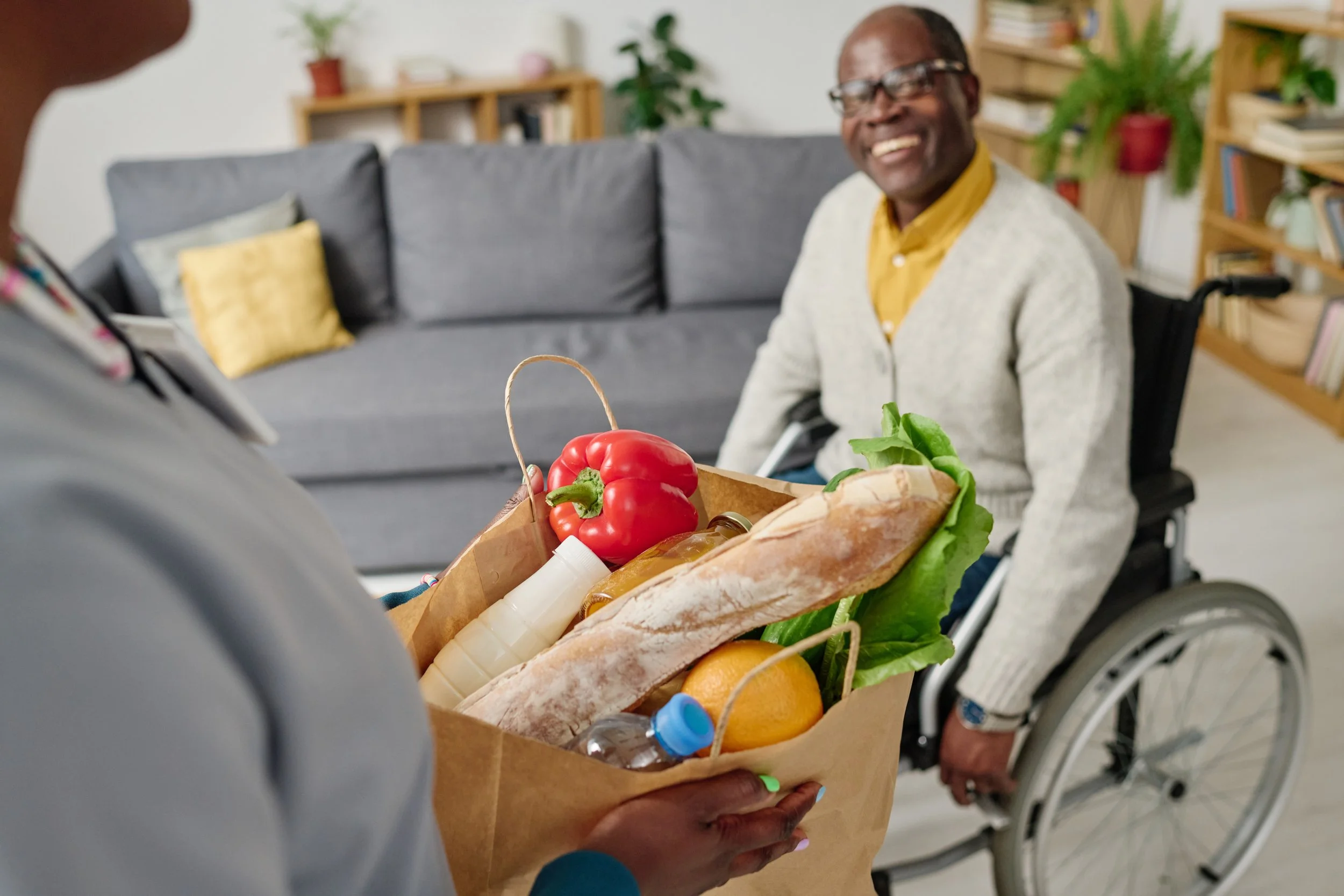 An elderly man in a wheelchair smiling and receiving a paper bag filled with groceries, including a loaf of bread, a red bell pepper, and lettuce, in a living room setting.