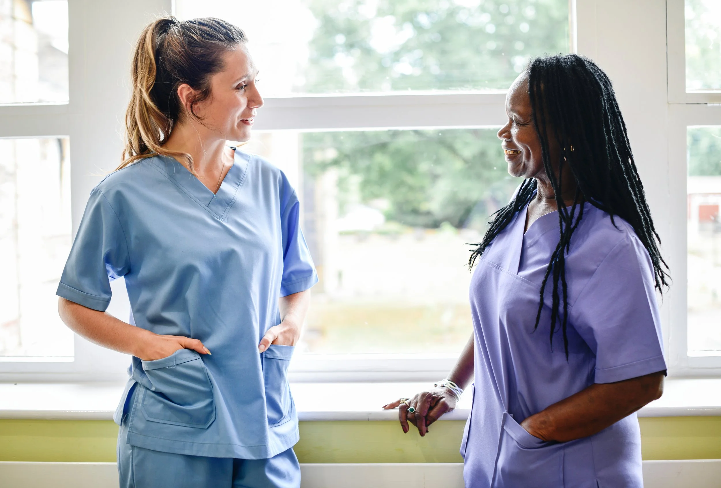 Two care providers in scrubs having a conversation in front of a window with greenery outside.