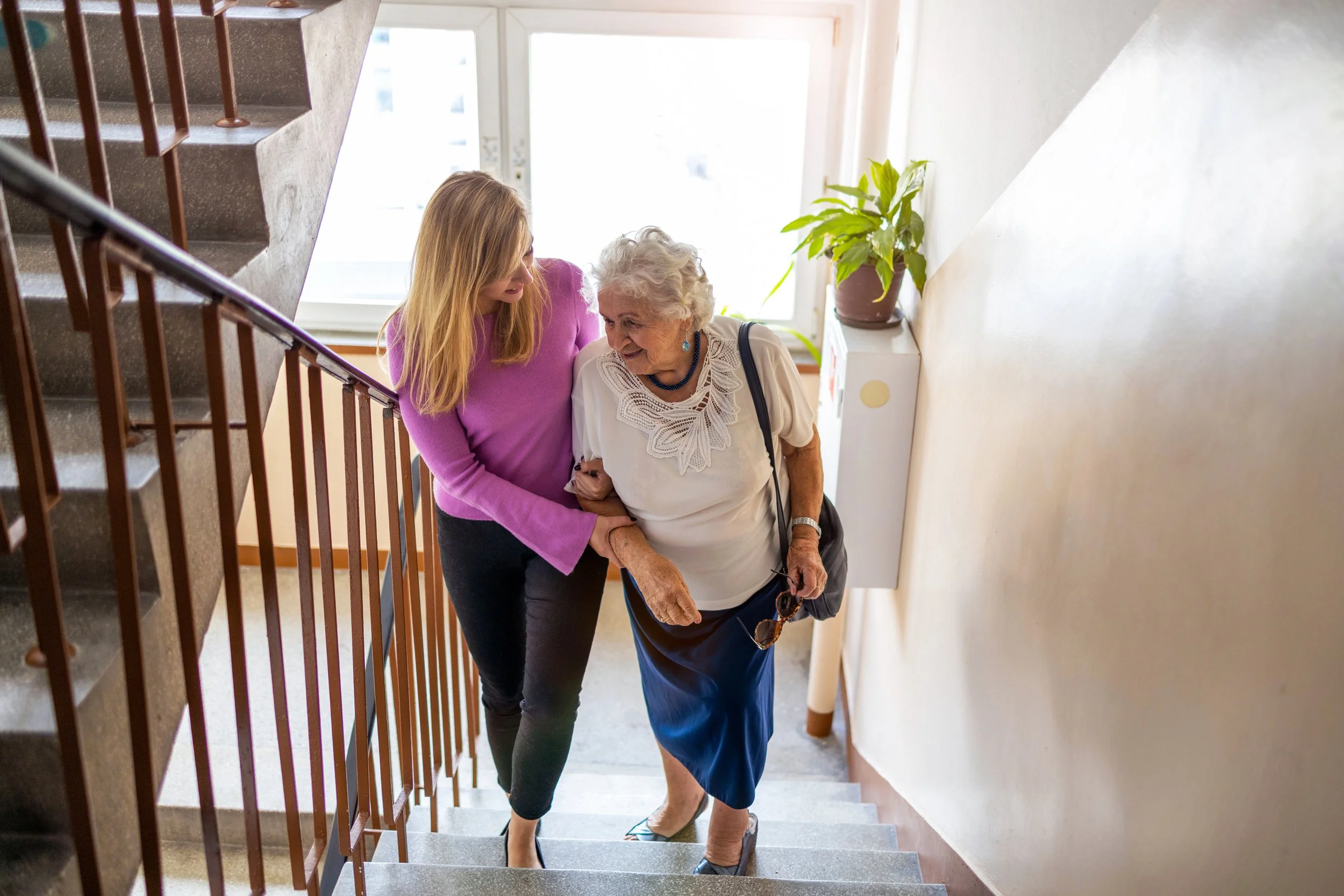 A young woman helping an elderly woman down a stairwell in a residential building.